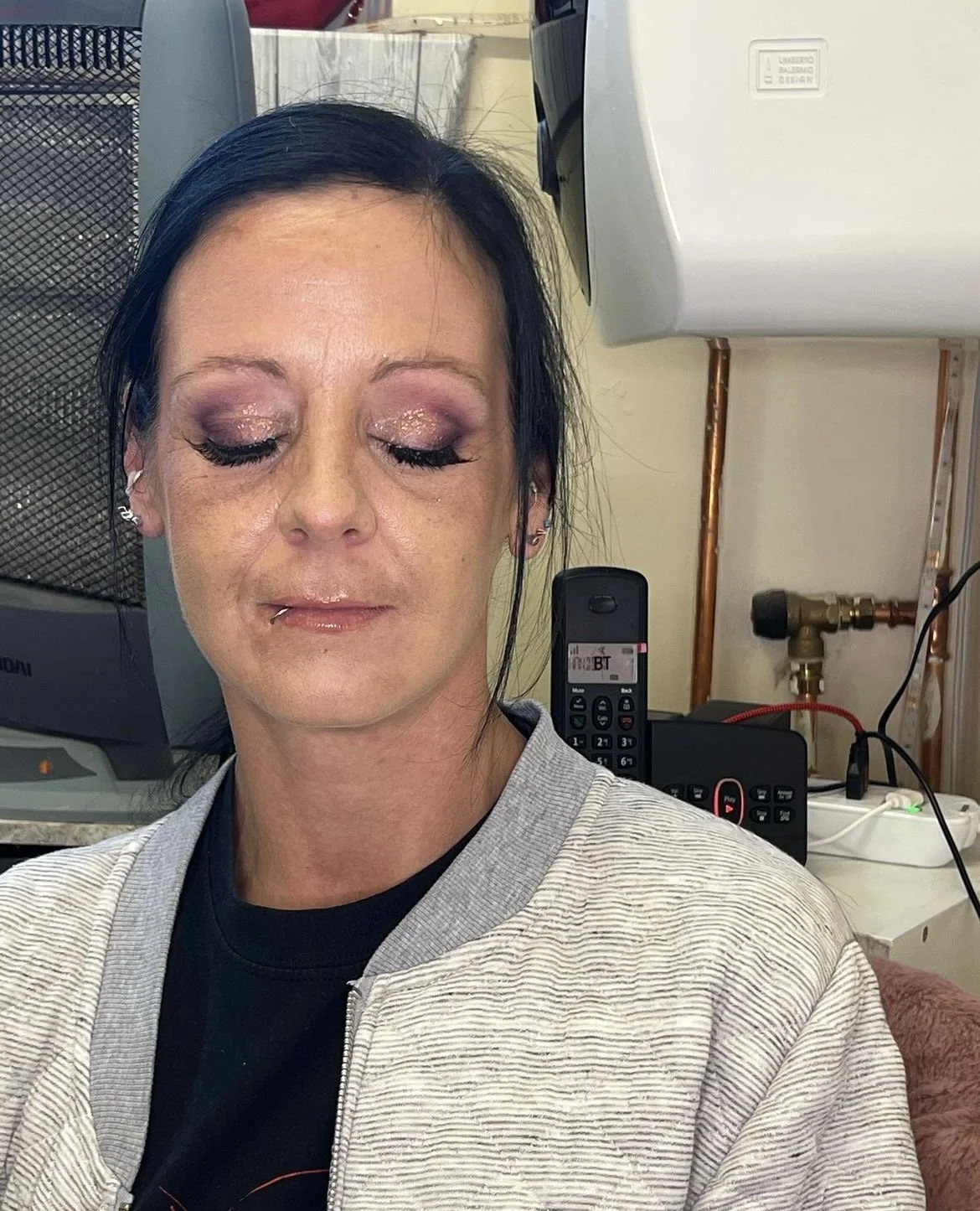A woman with dark hair, glittery eye makeup, and a lip piercing sitting with her eyes closed in a room with a landline phone, remote control, and various electrical cords in the background.