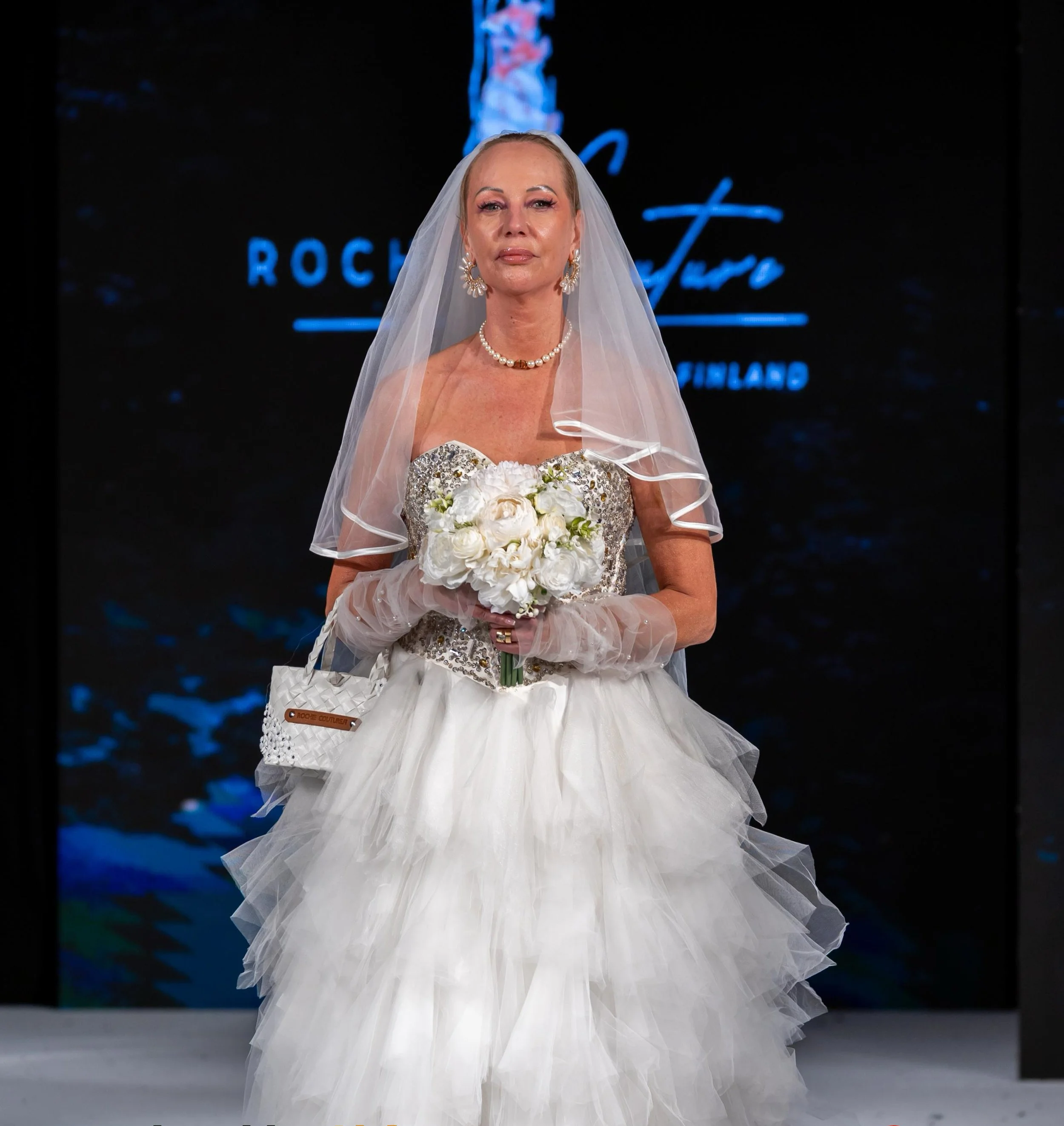 A woman wearing a wedding dress and veil holding a bouquet of white flowers on a runway.