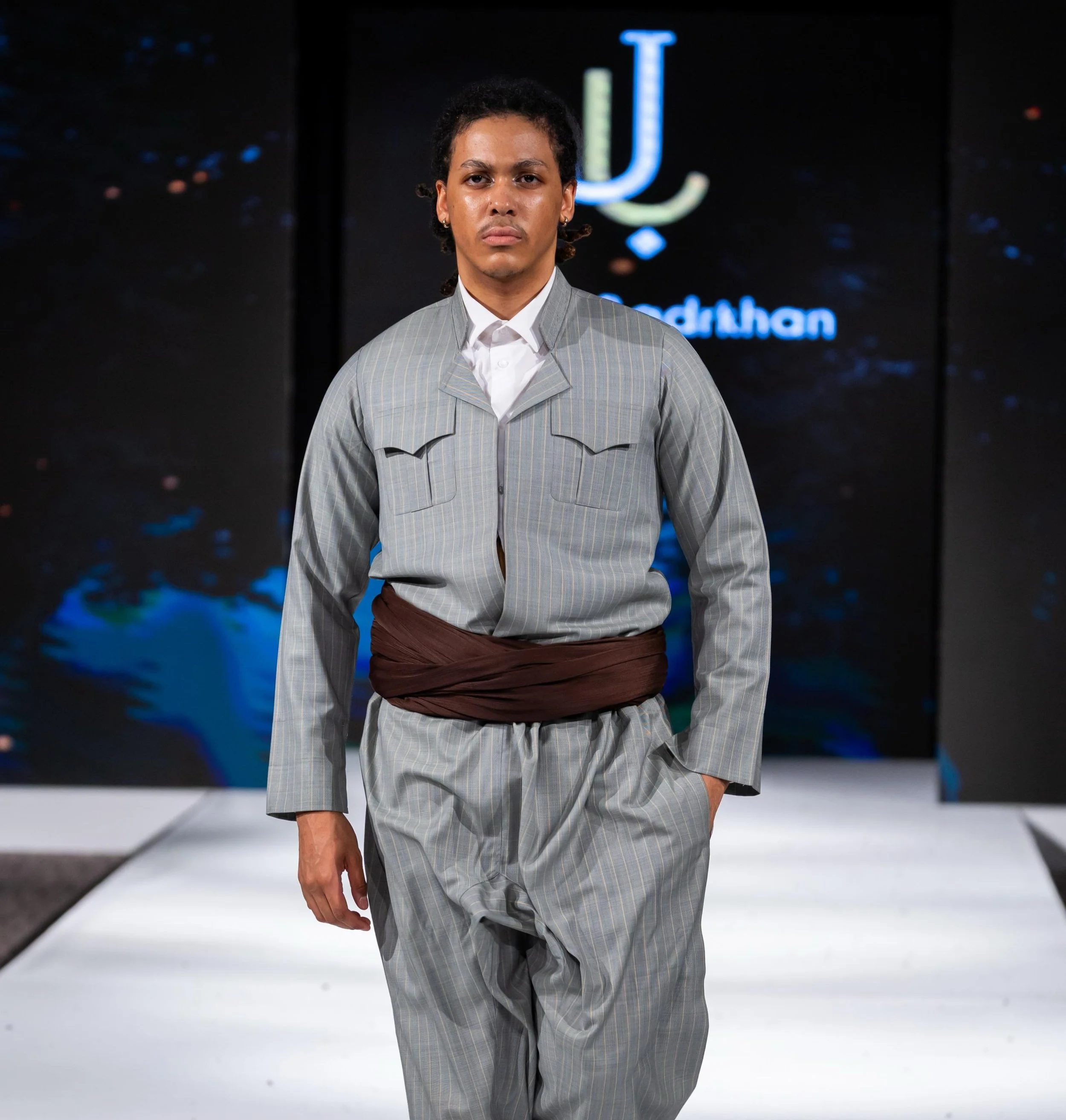 A young man walking down a runway during a fashion show, wearing a light gray striped suit with brown detailing and a white shirt underneath, with a dark background and a neon logo that reads 'j' in the background.