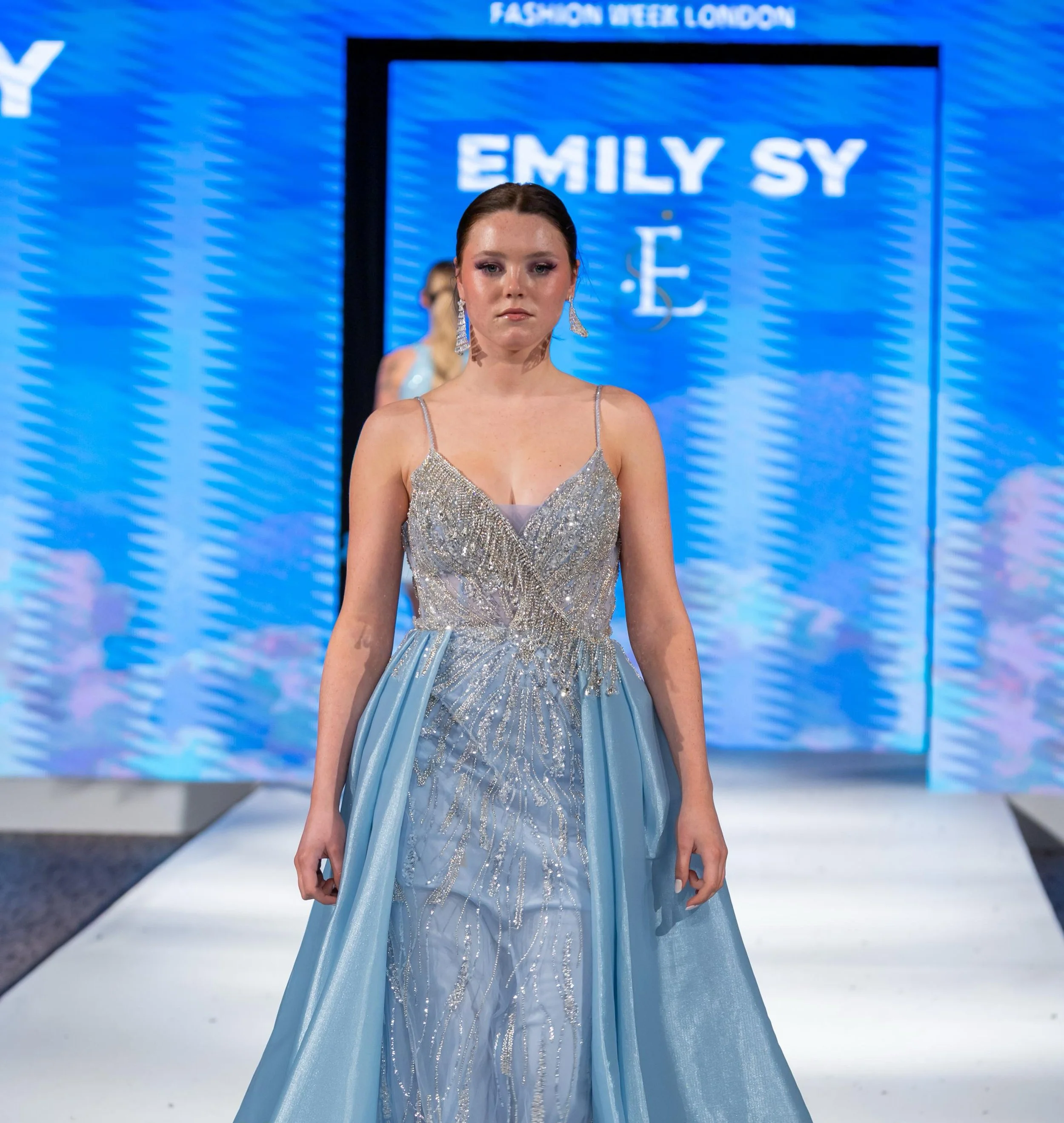 A model walking on the runway at Fashion Week London wearing a shimmering silver and blue gown with intricate beadwork and a flowing train.