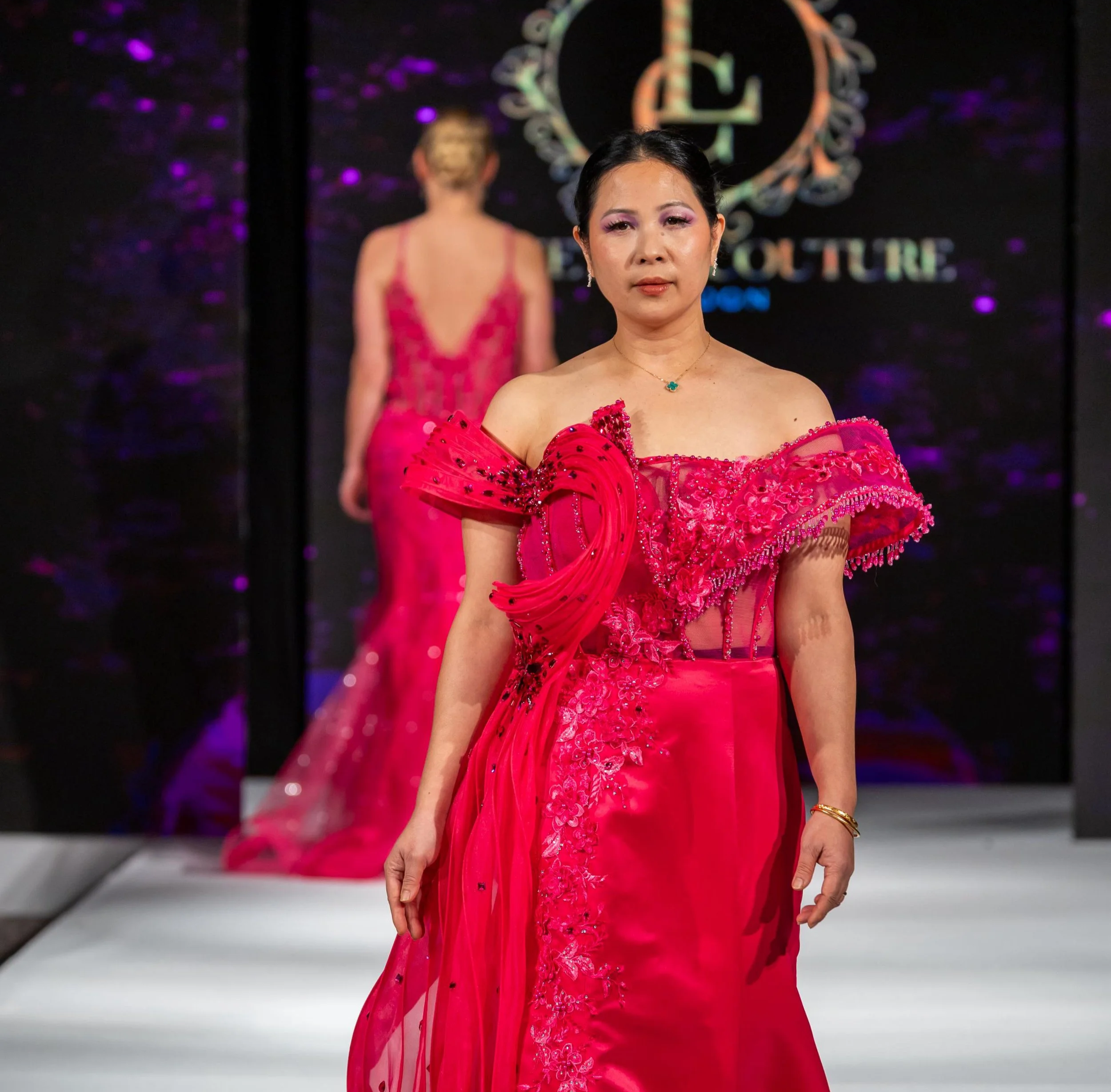 Model walking down runway in a bright red, intricately embroidered dress with off-the-shoulder puffed sleeves during a fashion show.