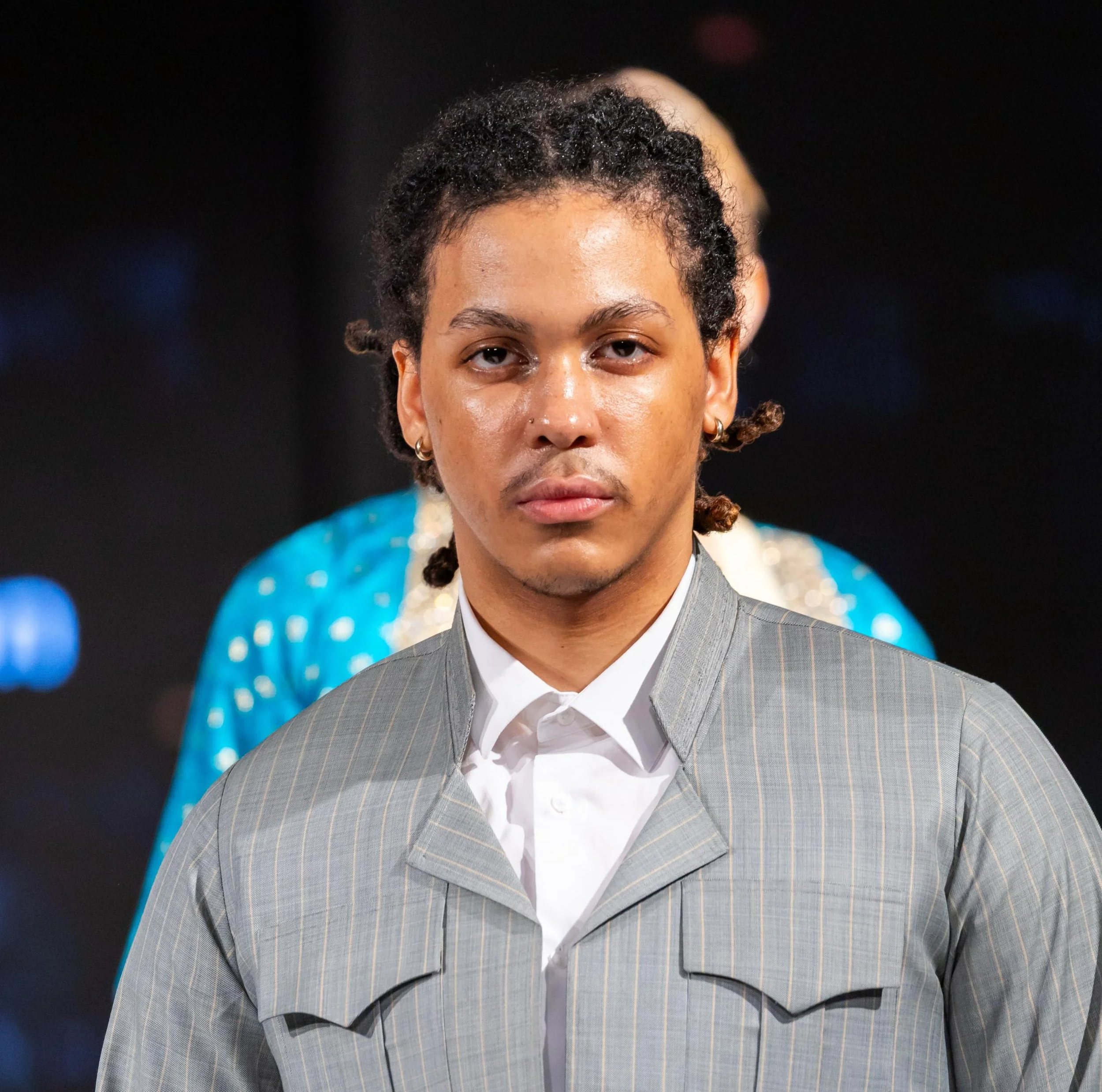 A person with short curly hair, wearing multiple earrings and a gray pinstripe suit with a white shirt, standing against a dark background with blue lights.