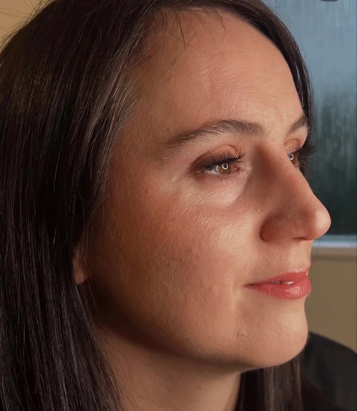 Close-up side profile of a woman with fair skin, brown hair, and brown eyes, looking out a window with rainy weather outside.