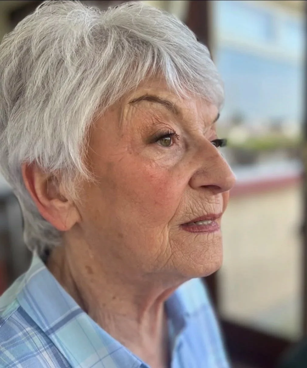 Close-up of an elderly woman with short gray hair, wearing a blue plaid shirt, looking out a window with a thoughtful expression.