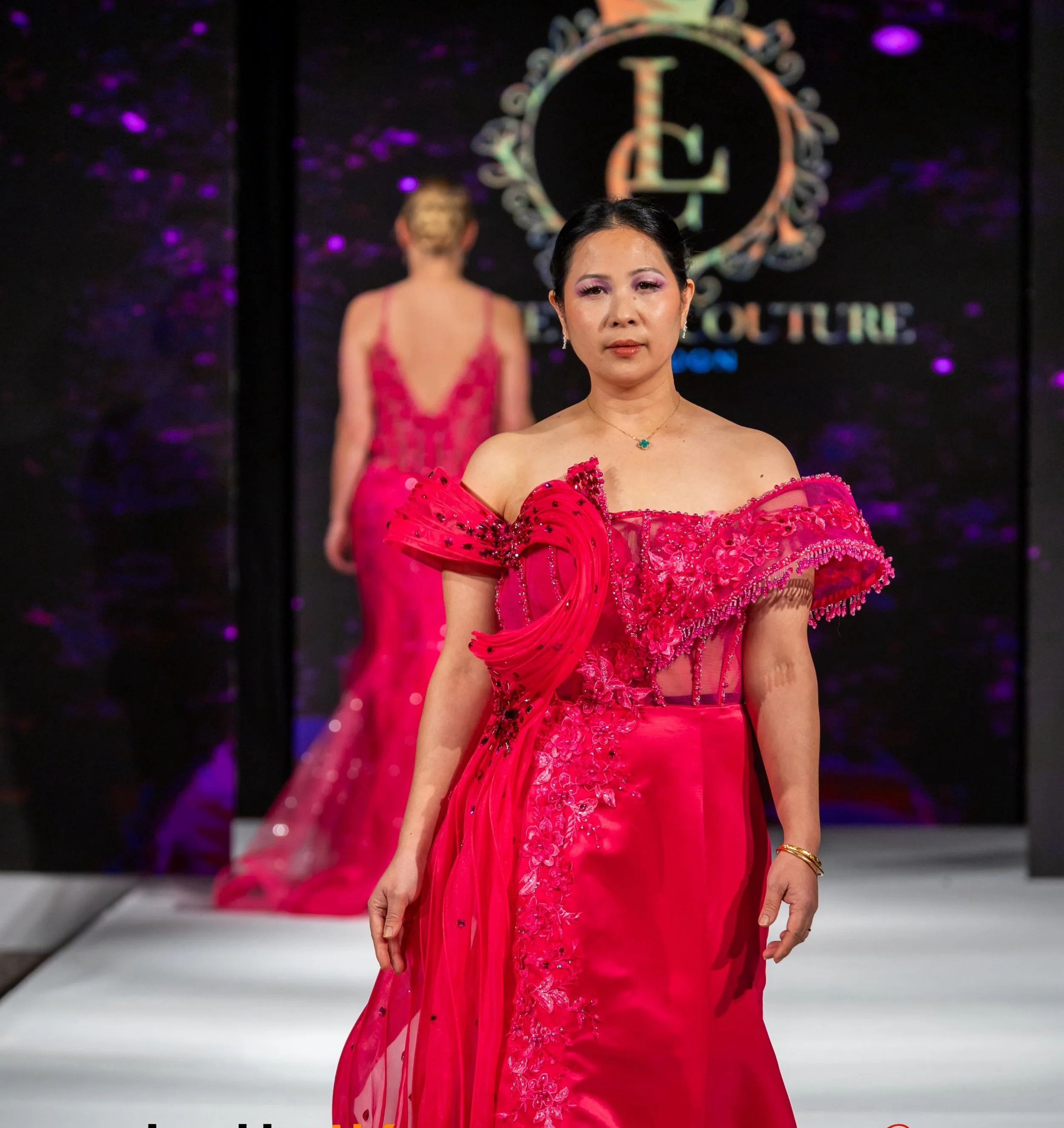 A woman wearing a bright pink dress with floral embroidery and sheer sleeves walks down a runway. She has dark hair styled back, with makeup emphasizing her eyes, and wears a necklace and earrings. Behind her, a mirror or reflective surface shows her