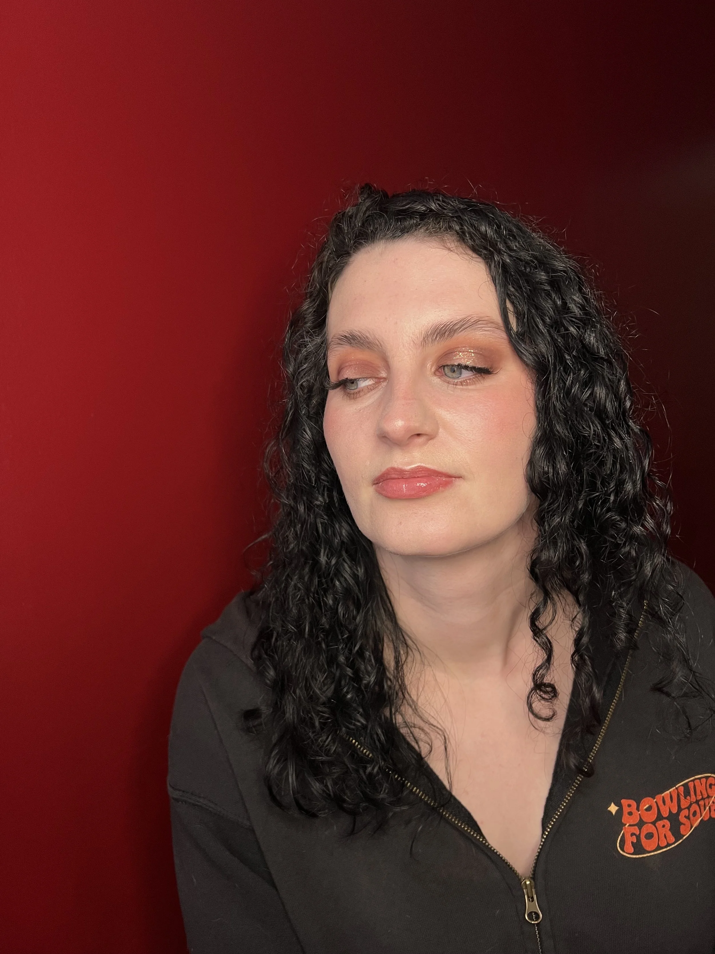 A woman with black curly hair and light makeup, wearing a black zip-up hoodie with a logo that says "Bowling for Souls," standing against a red background.