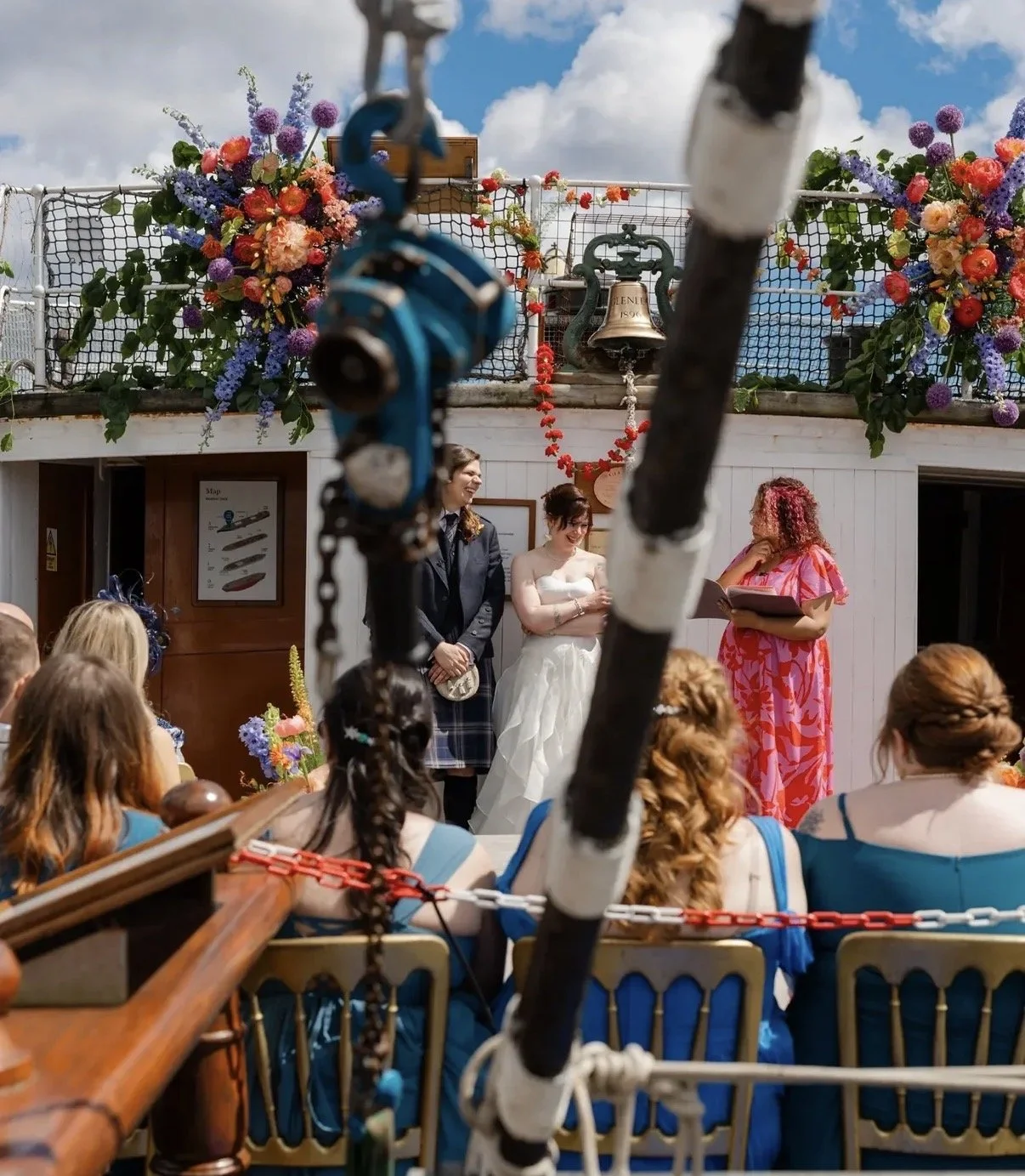 A wedding ceremony on a ship's deck with flowers and decorations. The bride and groom stand with an officiant as guests watch. The scene is outdoors with blue sky and clouds.