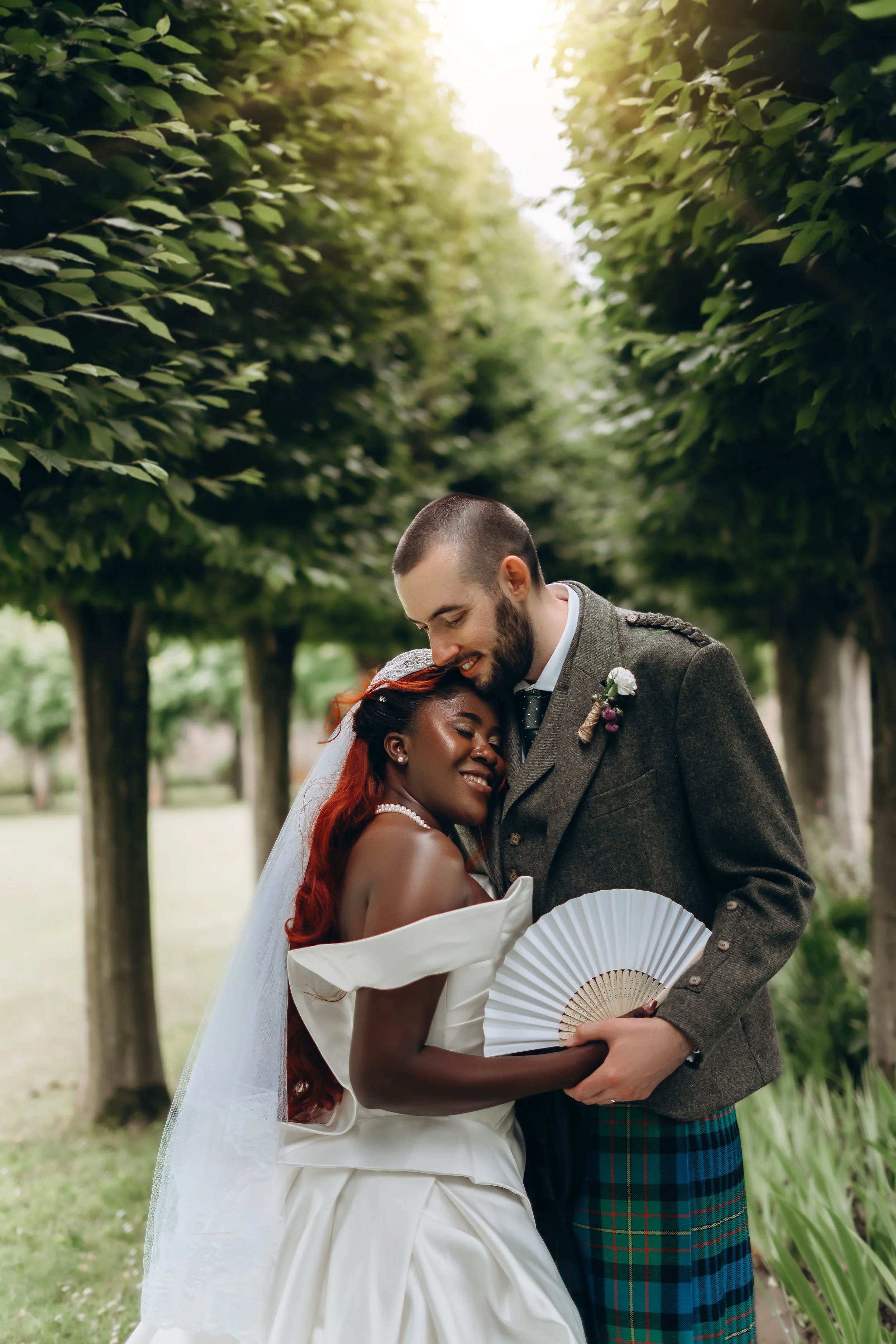 A happy couple, a woman with long red hair and a man with a beard dressed in a Scottish kilt and jacket, sharing an embrace outdoors with trees and sunlight in the background.