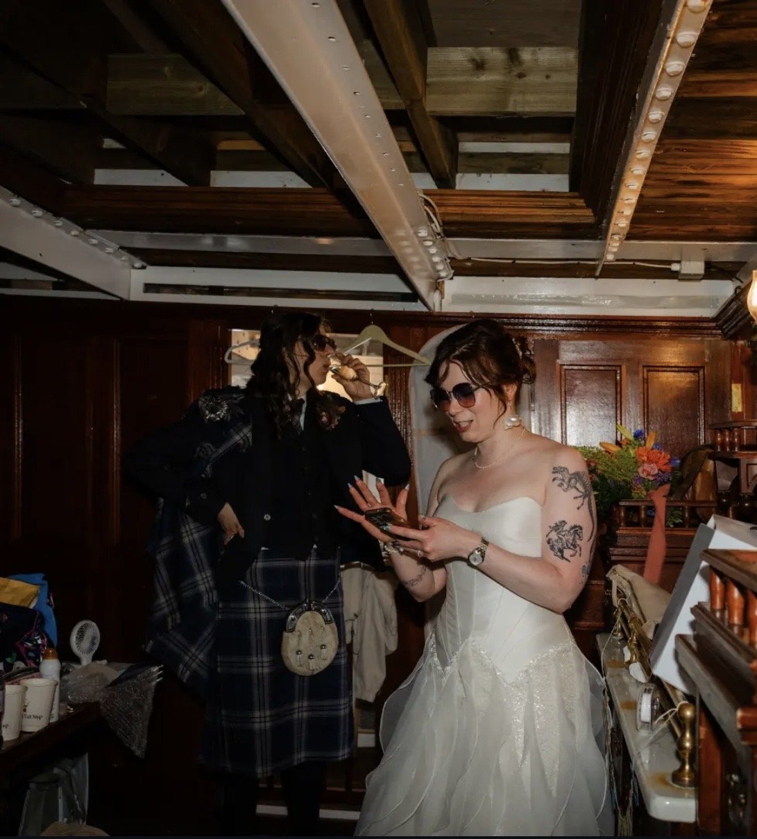 Two women, one in a wedding dress and the other in a tuxedo, are inside a room with wooden walls and ceiling. The woman in the wedding dress is holding a phone, while the other woman is wearing sunglasses and taking a sip from a glass. There are flow