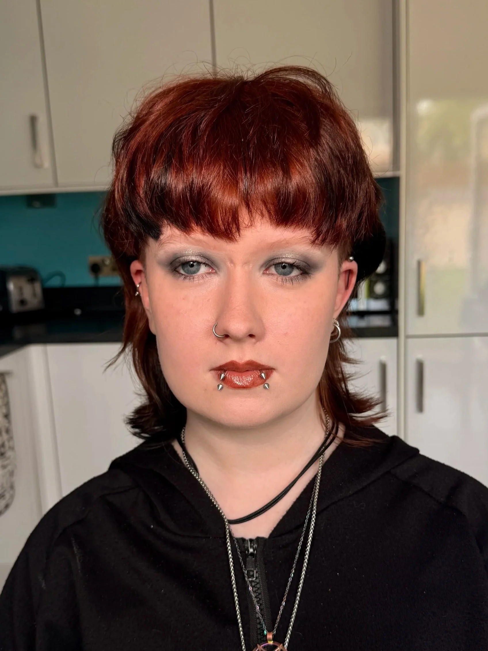 Close-up of a person with short red hair, pale skin, blue eyes, silver makeup, nose piercing, and lip piercings, wearing silver necklaces, and a black zip-up hoodie, in a room with cabinets in the background.