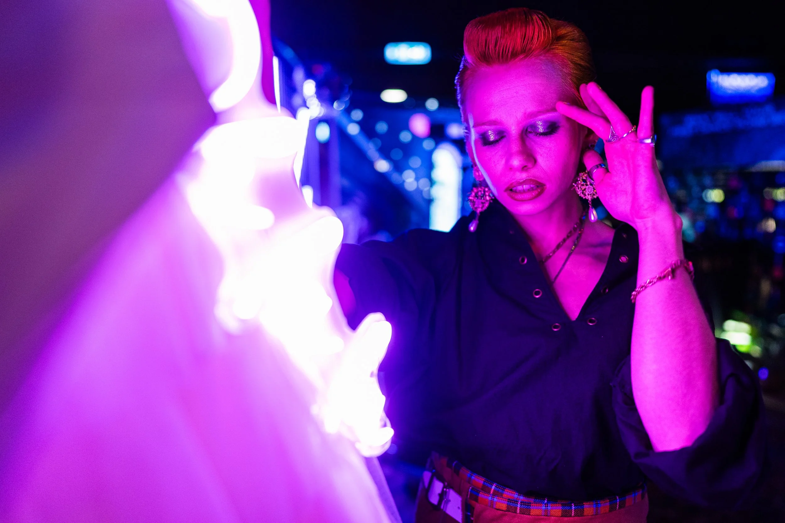 A person with brightly colored hair and elaborate jewelry is in a dark room illuminated by vibrant neon purple and blue lights, with their eyes closed and one hand near their face, creating an intense atmosphere.