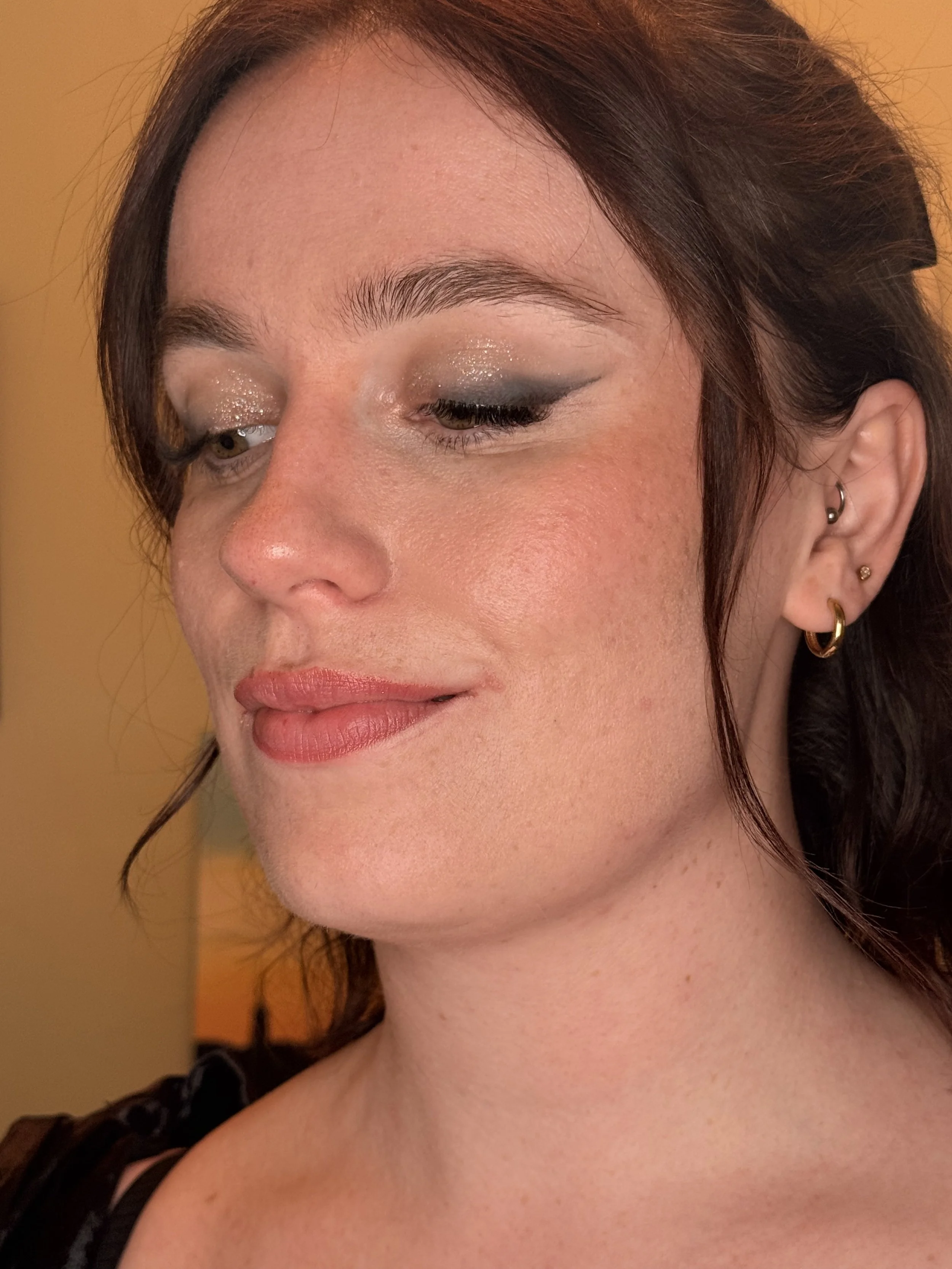 A close-up of a woman's face with makeup, showing her closed eyes, earrings, and part of her shoulder, with a warm-colored background.