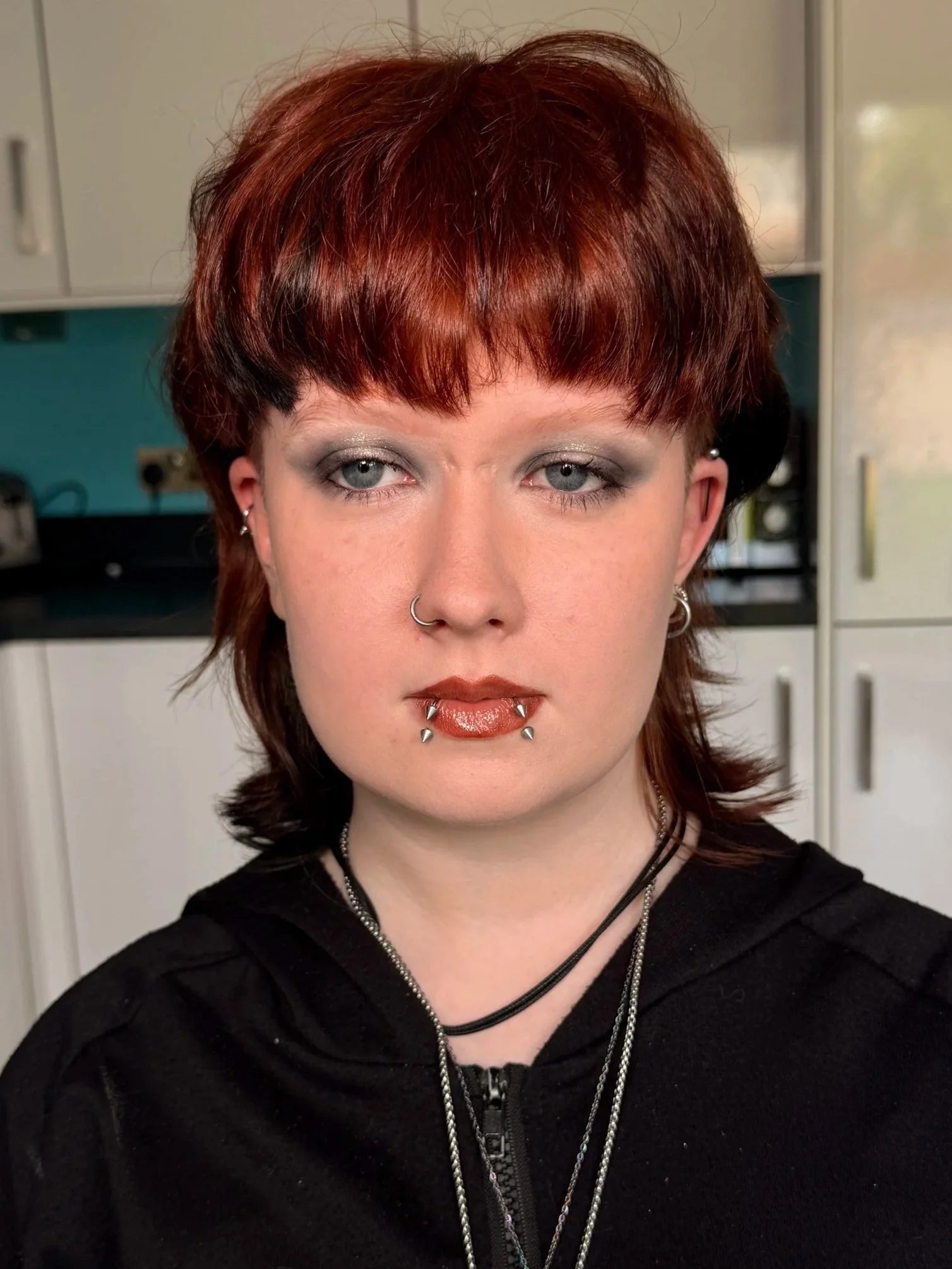 Close-up of a woman with short red hair, pale skin, and multiple facial piercings, wearing dark makeup and jewelry, in a room with cabinets in the background.