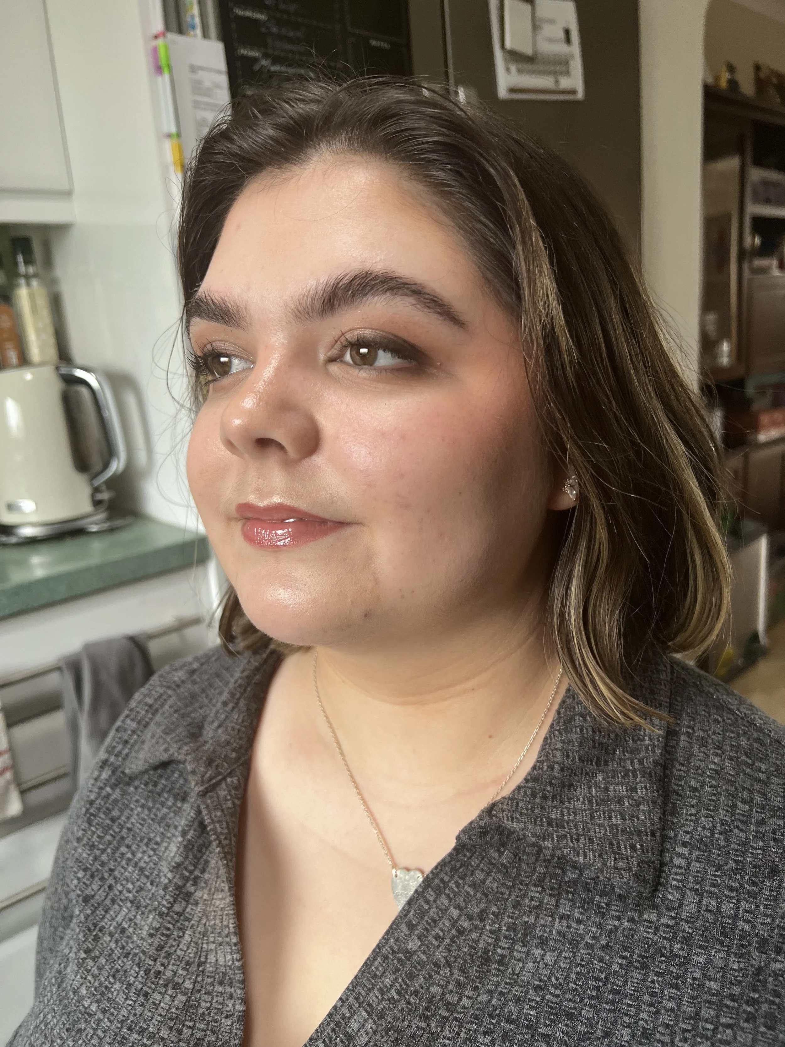 A close-up of a young woman with brown hair, wearing makeup, a gray textured blazer, a necklace, and pearl earrings, standing in a kitchen with appliances and shelves in the background.