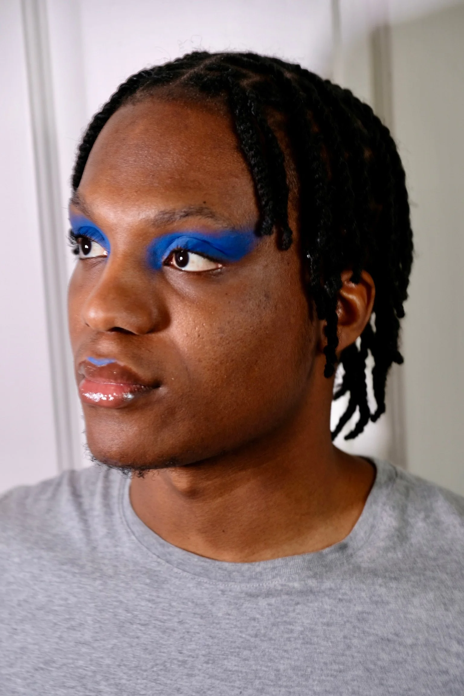 A person with dark skin and braided hair with twists, wearing bright blue eyeshadow and gloss on lips, dressed in a gray t-shirt, looking slightly to the side.