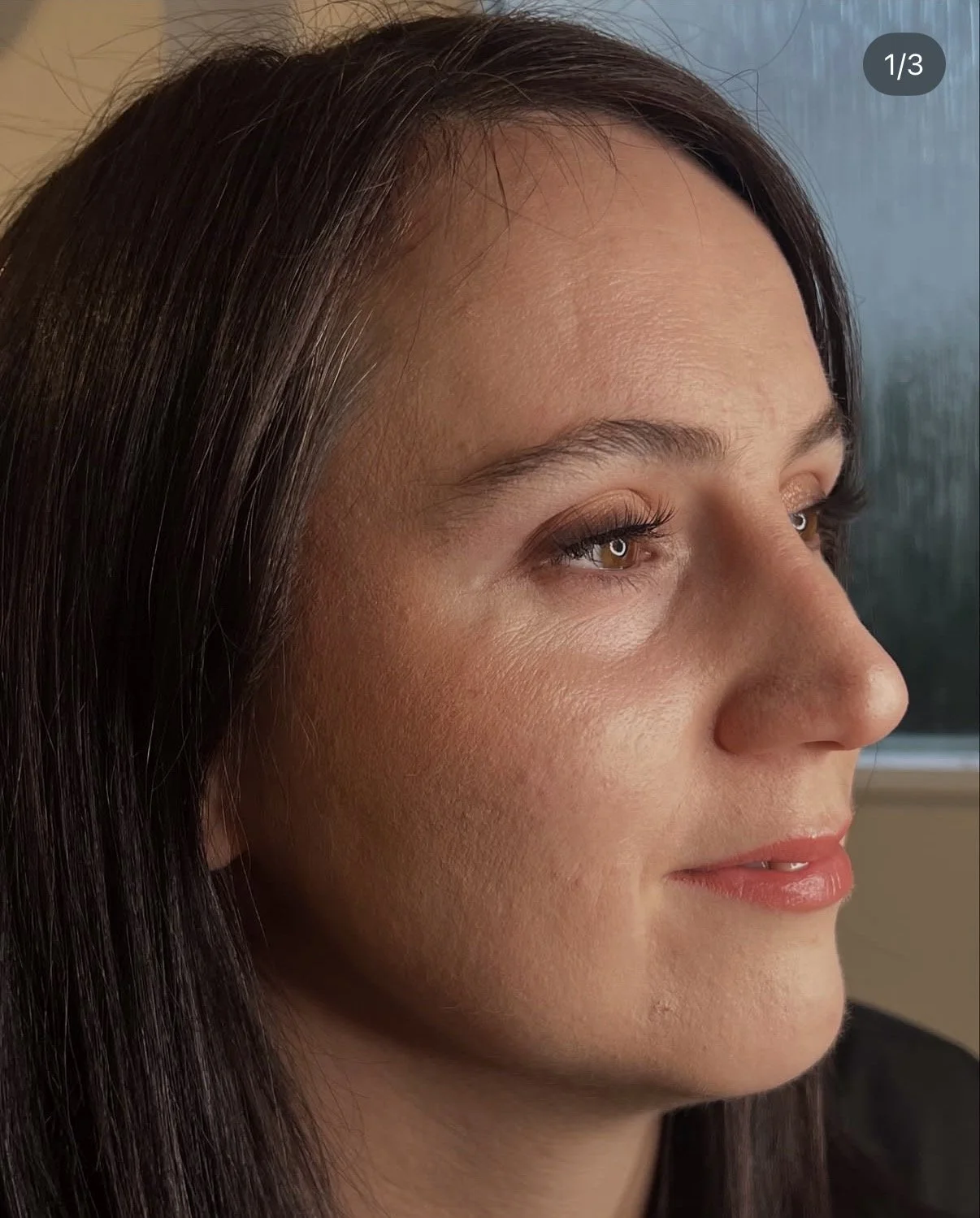 Close-up of a woman's face in profile with brown hair, brown eyes, and natural makeup, indoors by a window.