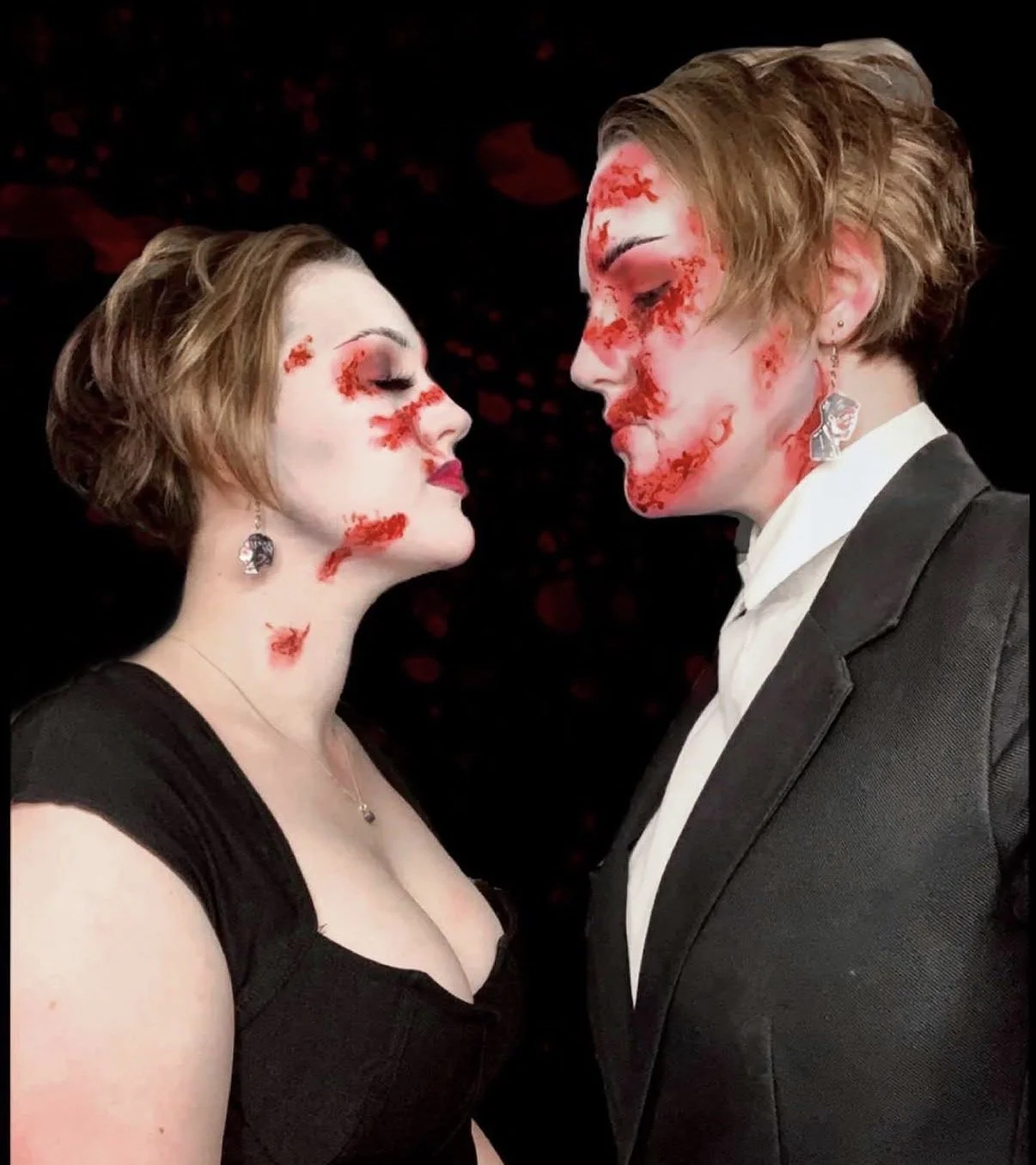 A man and woman face each other with closed eyes, both with makeup and fake blood on their faces, suggesting a Halloween or horror theme. The woman is wearing a black dress and the man is in a suit.