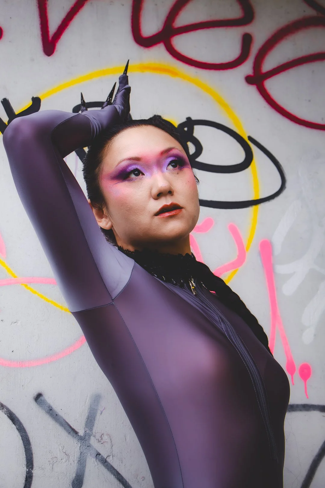 A person with bold purple eye makeup and dark lipstick posing against a wall with colorful graffiti. They are wearing a tight-fitting purple and gray jacket, with one arm raised above their head and holding their hair back.