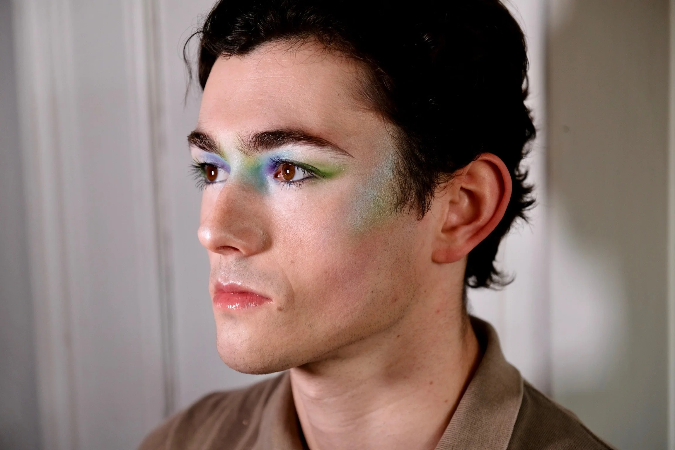 Close-up of a person with short black hair and colorful eye makeup, wearing a brown shirt, standing in front of a plain wall.