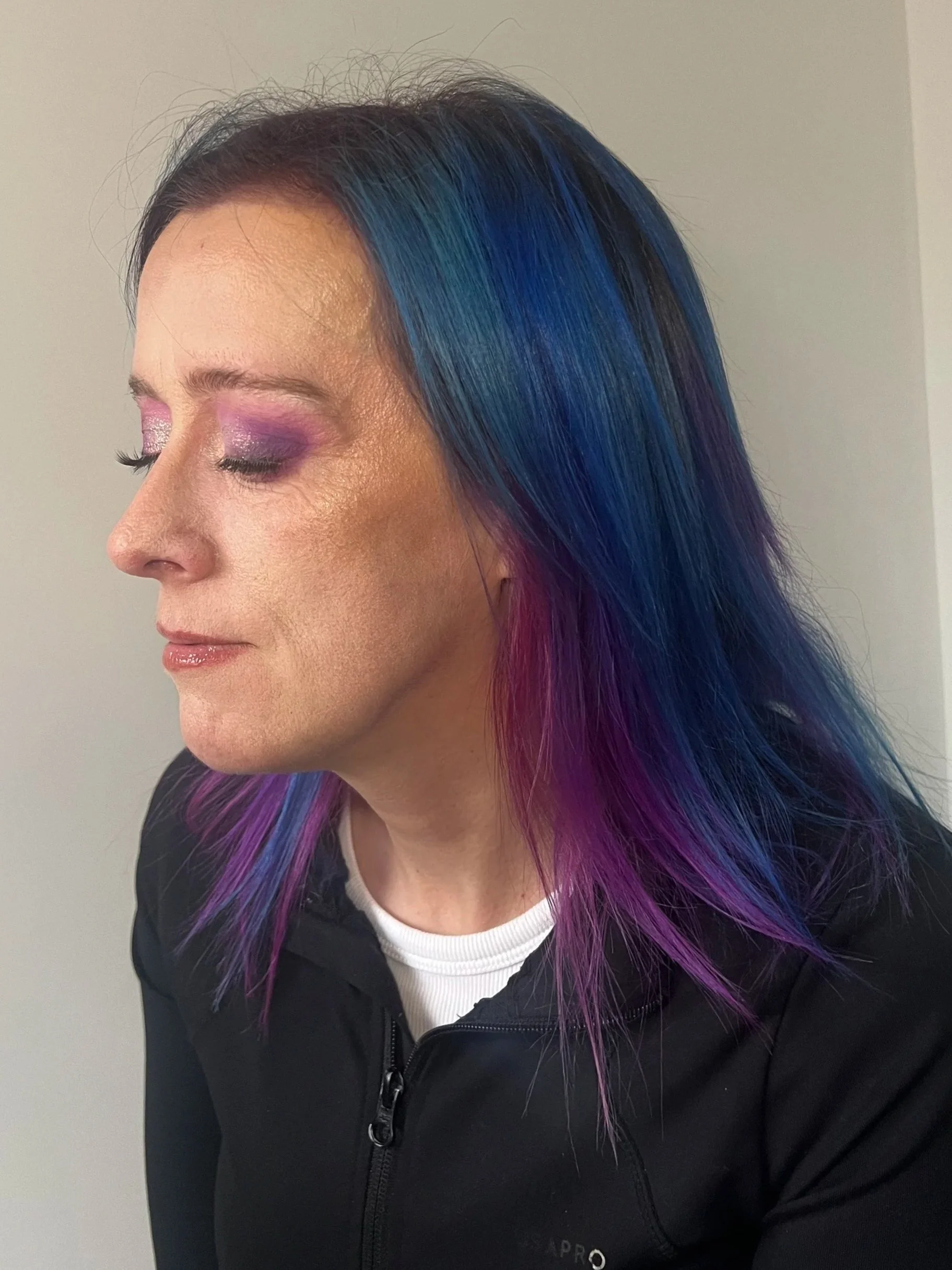 Woman with black hair dyed in vibrant purple, blue, and green colors, wearing a black jacket and white shirt, with makeup including purple eyeshadow, posing with her eyes closed.