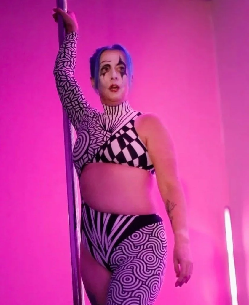 A woman with blue hair and clown makeup performing on a pole in a room with pink lighting, wearing a black and white geometric patterned outfit.