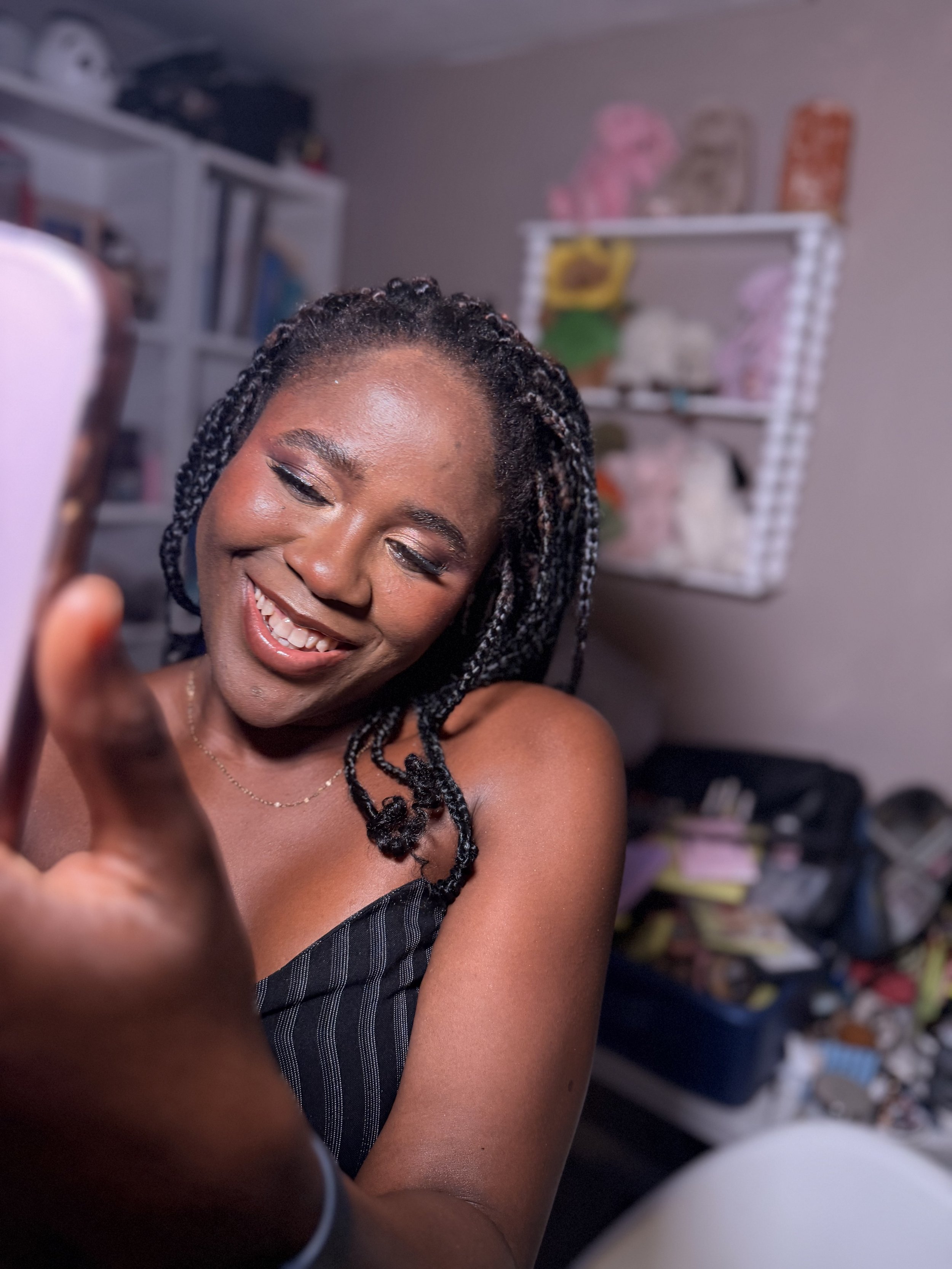 A smiling woman with braided hair taking a selfie in a room with shelves filled with colorful packages and items in the background.