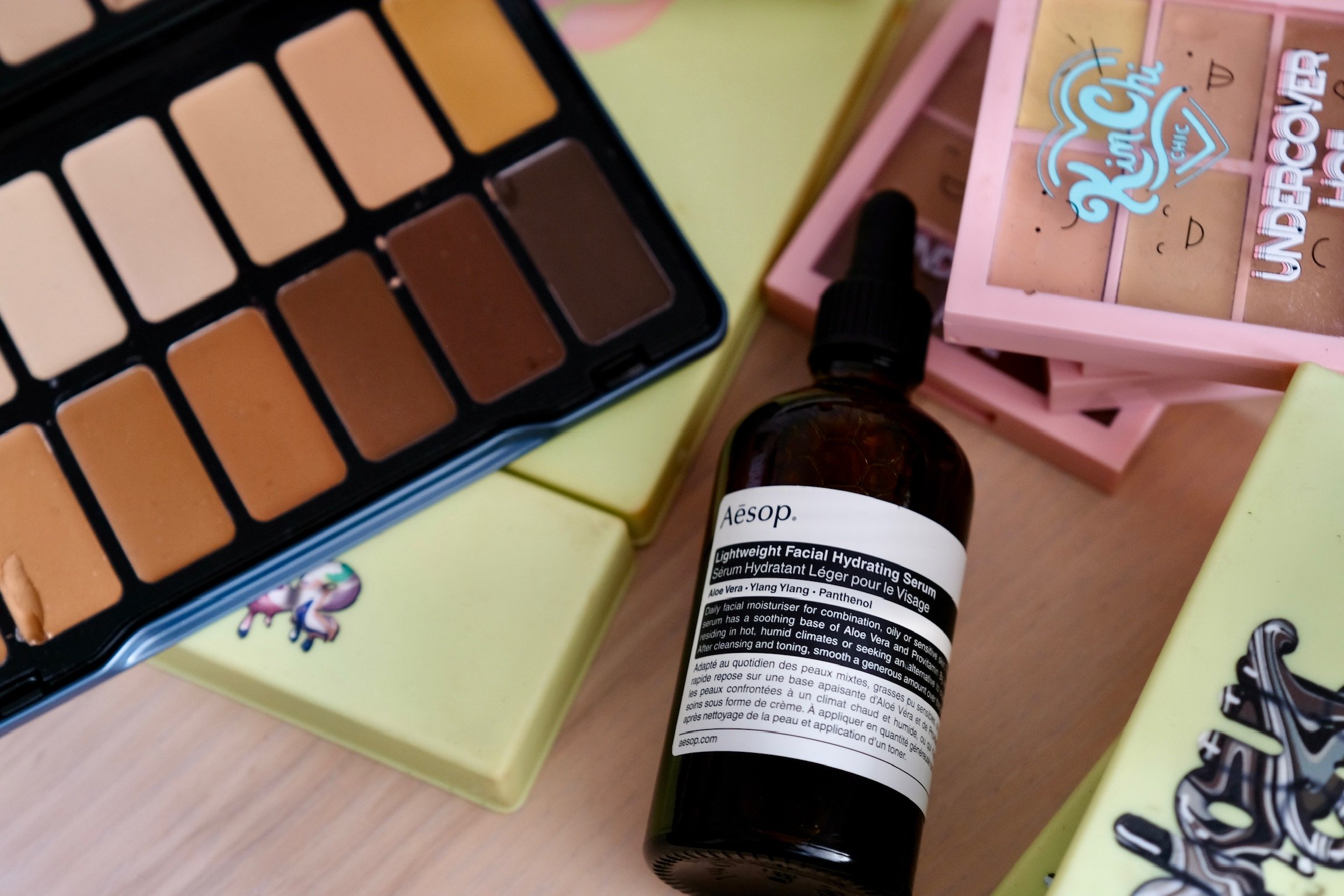 A skincare bottle labeled Aesop Lightweight Facial Hydrating Serum with aloe vera, ylang ylang, and panthenol, placed among makeup and skincare products.