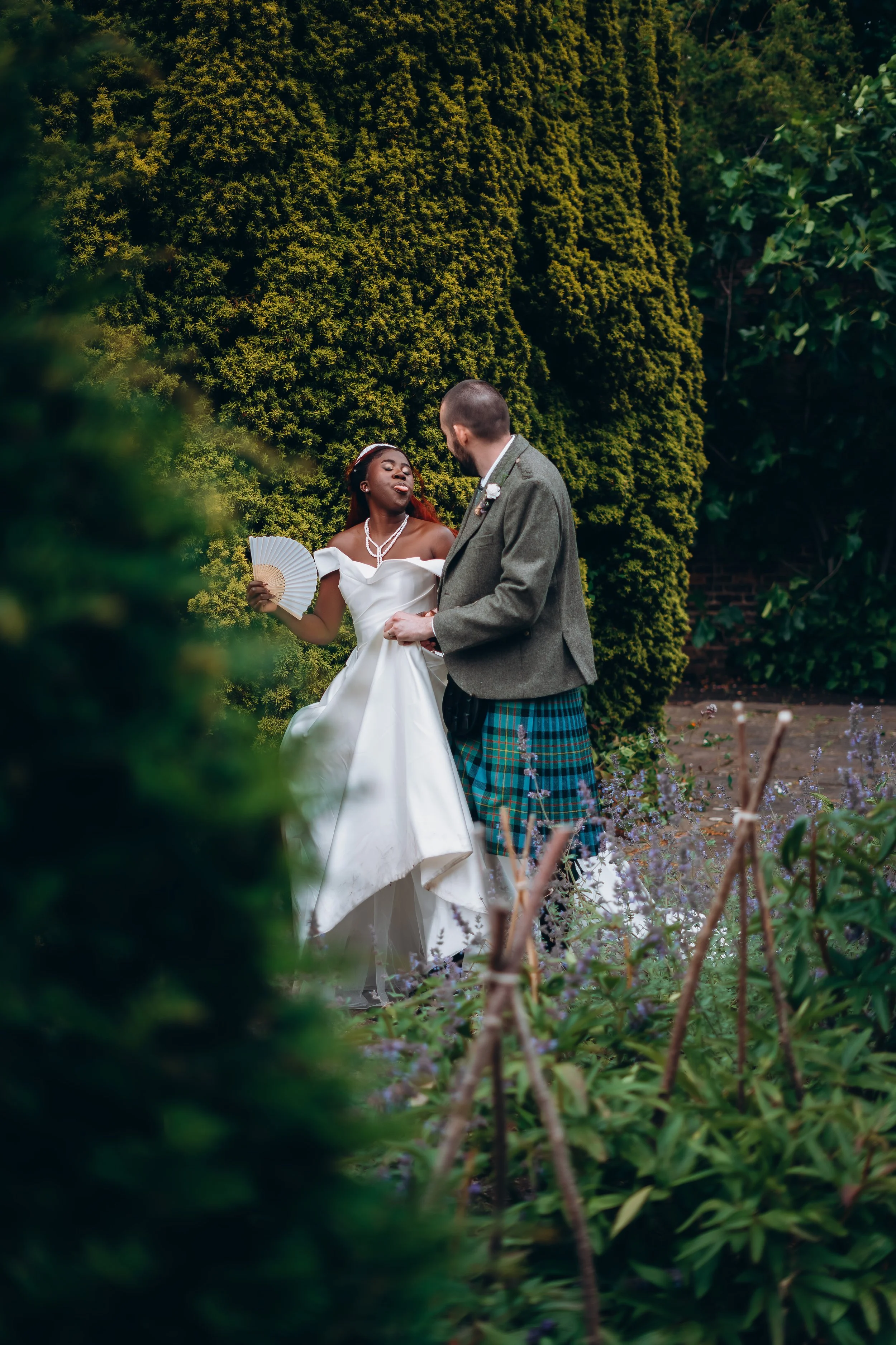 A bride in a white wedding gown holding a fan deals with a groom wearing a kilt, in a garden setting with green foliage.