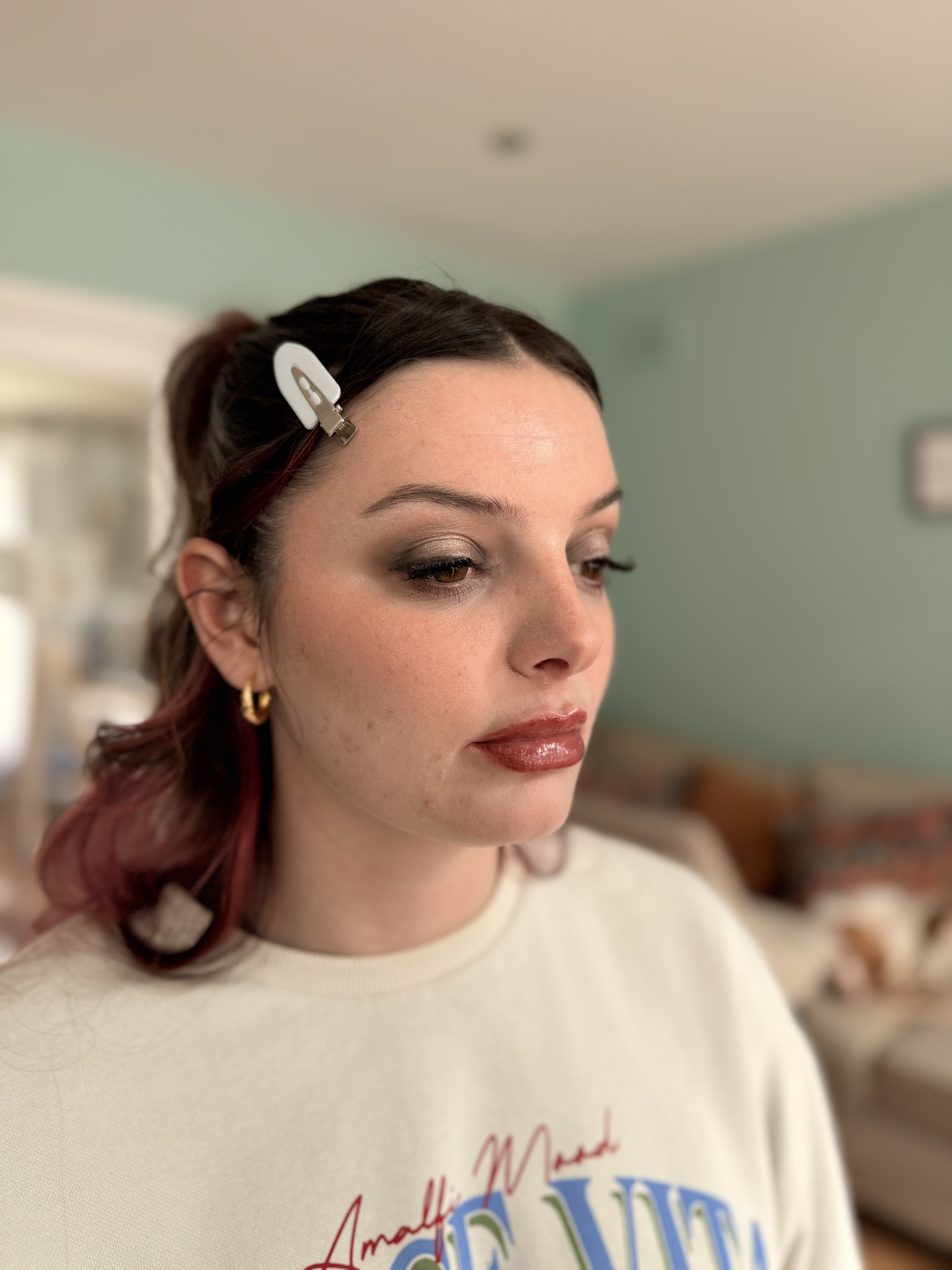 A woman with dark brown and red hair, secured with a white hair clip, is looking down slightly. She has makeup on, including eyeshadow, mascara, and lipstick. She is wearing a white sweatshirt with colorful text and appears to be indoors, possibly in