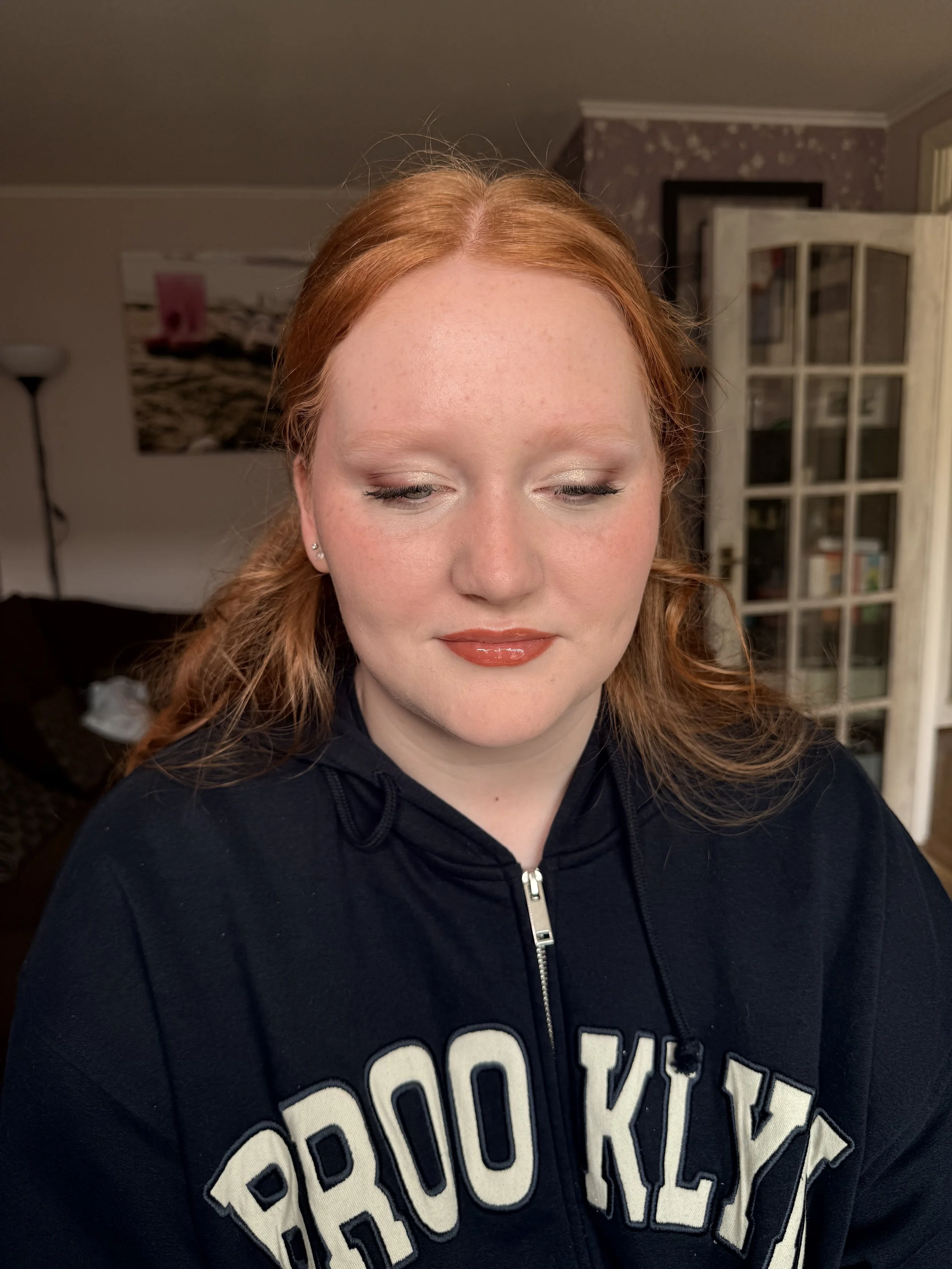 A woman with red hair and fair skin wearing makeup, a black hoodie with large white letters, standing in a well-lit room with a bookcase and a painting in the background.