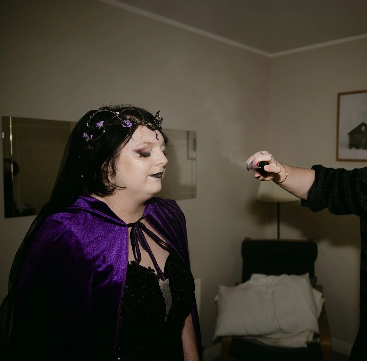A woman with black hair, makeup, and a purple velvet cloak, getting spray makeup applied in a room with beige walls and a bed.