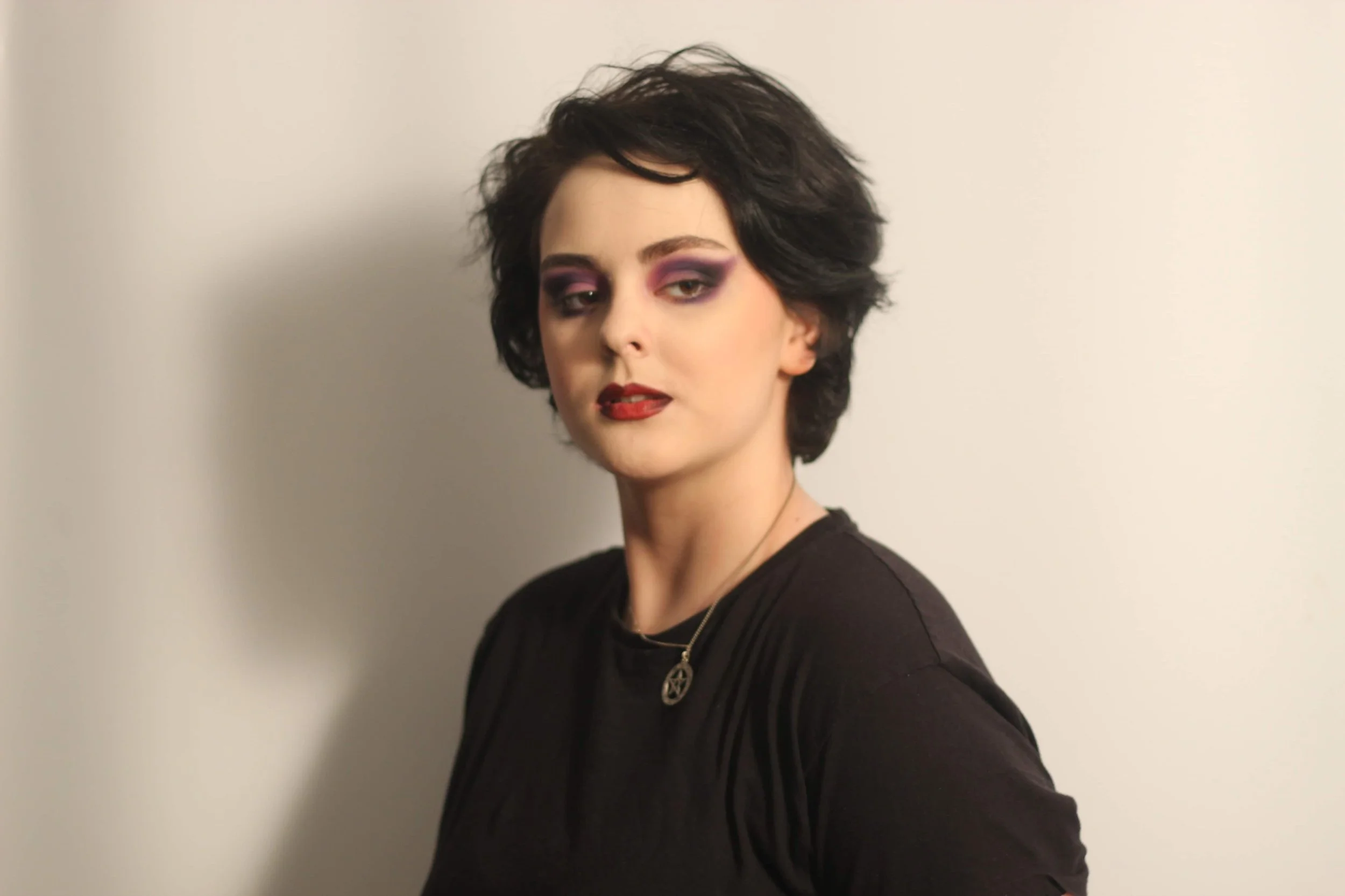 A woman with short dark hair, purple eyeshadow, red lipstick, wearing a black top, a pendant necklace with a pentagram, and standing against a plain light-colored wall.