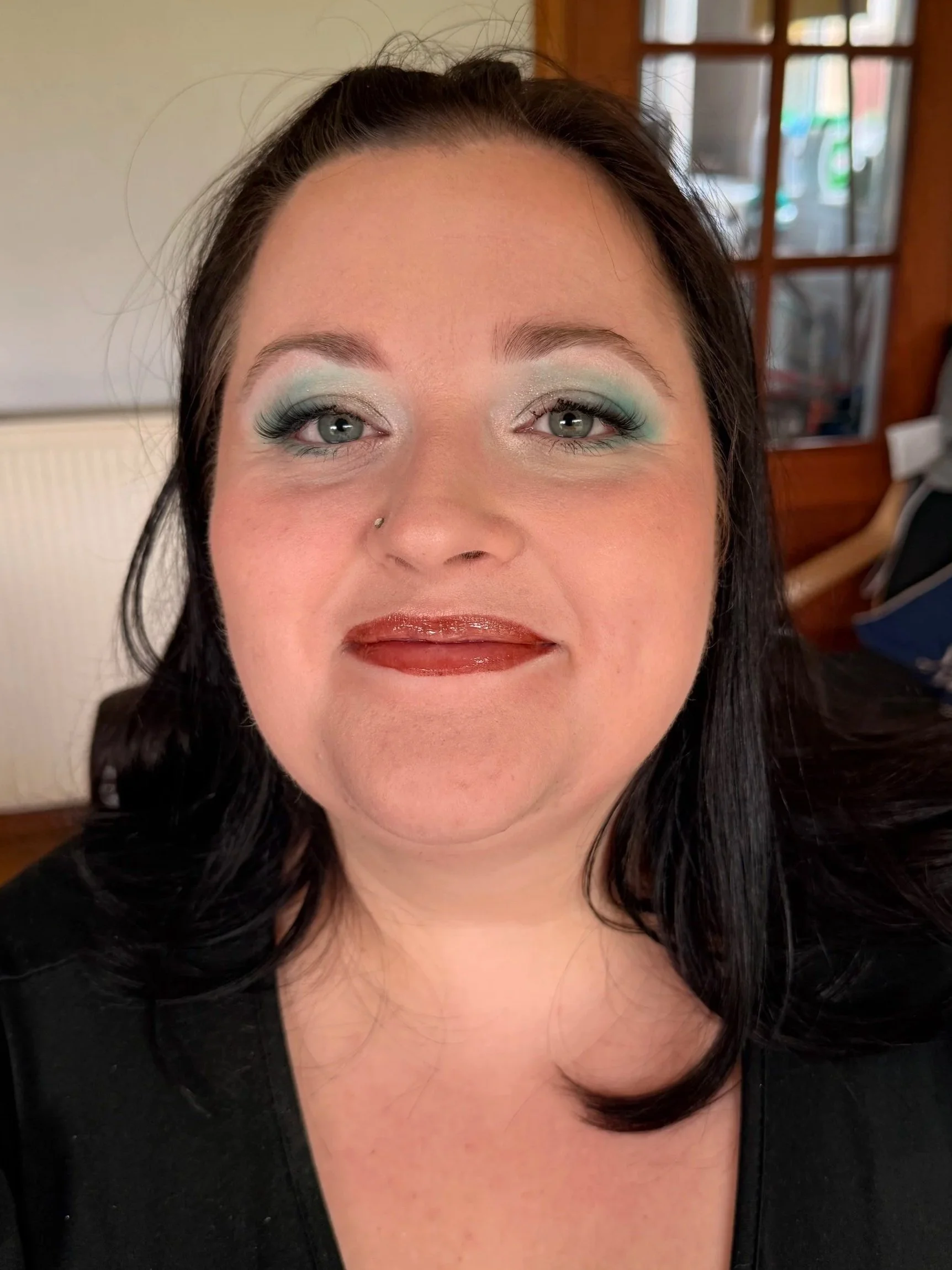 A woman with dark hair, wearing eye makeup with shades of blue and green, smiling slightly, wearing a black top, with a background showing a wooden door and glass window.
