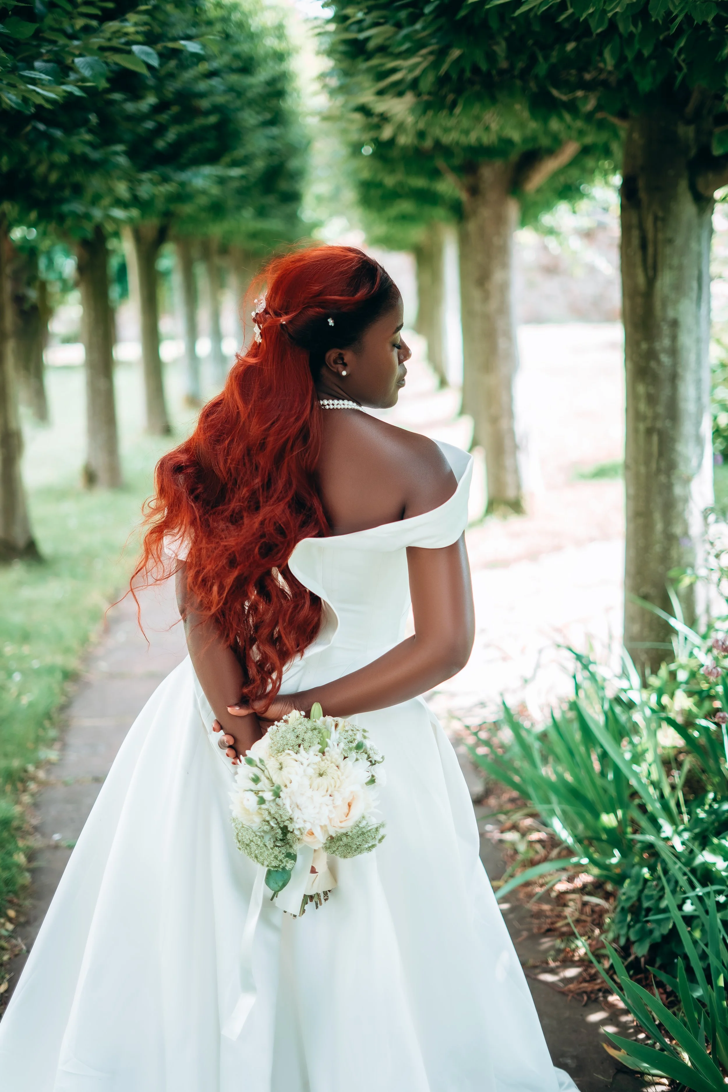 A woman with long, wavy red hair in a white off-shoulder dress holding a bouquet of flowers, standing in a tree-lined path.
