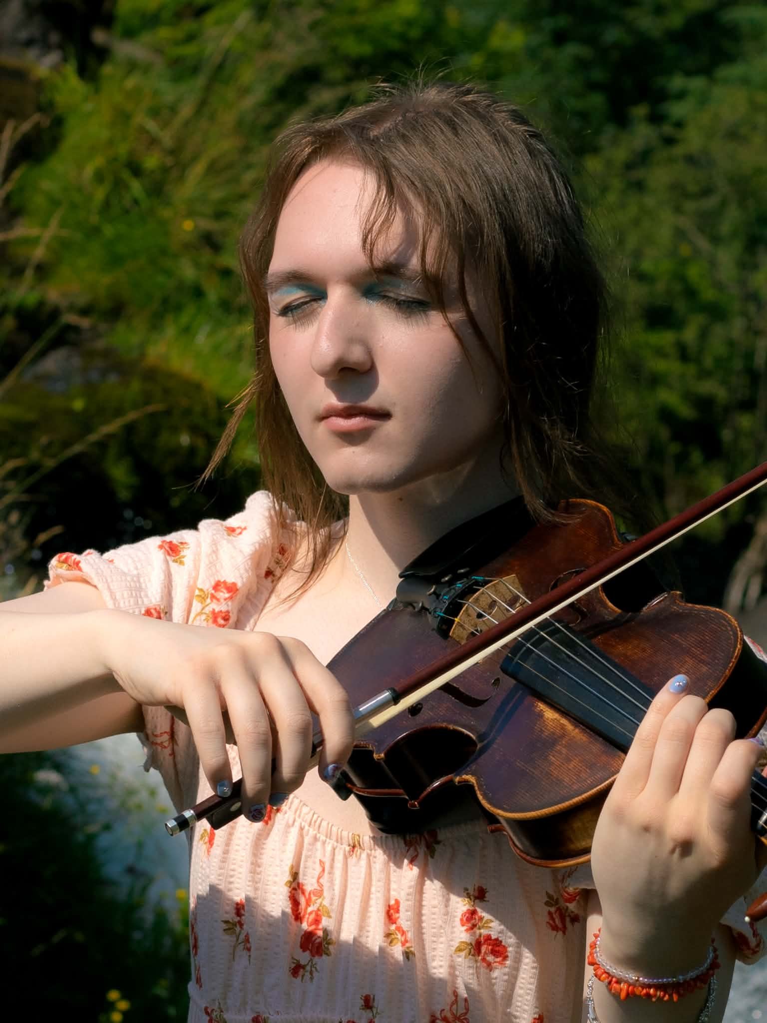 A young woman playing the violin outdoors in a green, wooded area, with sunlight highlighting her face and hands.