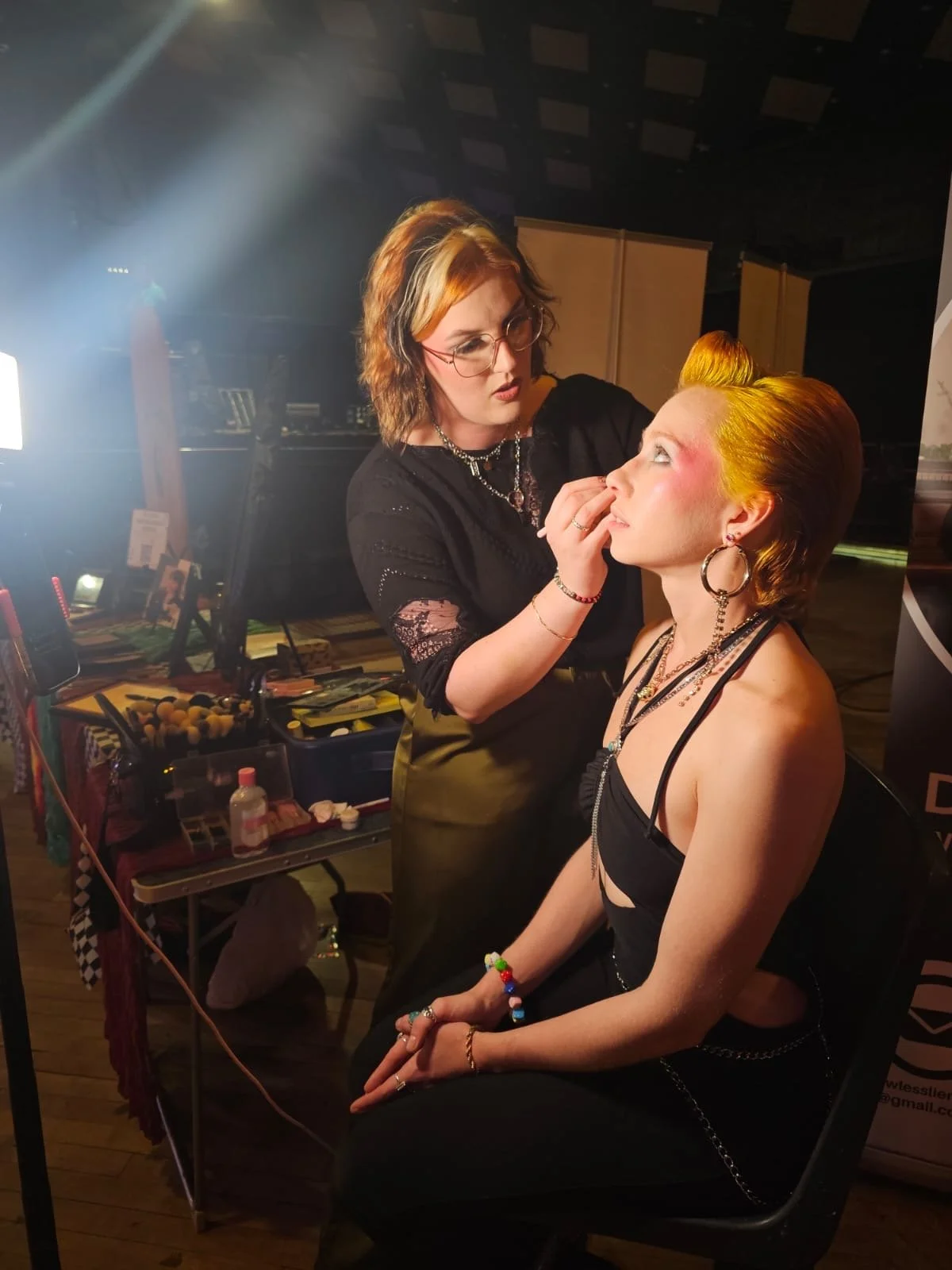 Makeup artist applies makeup to a sitting model with bright yellow hair, wearing earrings and a black top, in a backstage setting with makeup tools and supplies in the background.