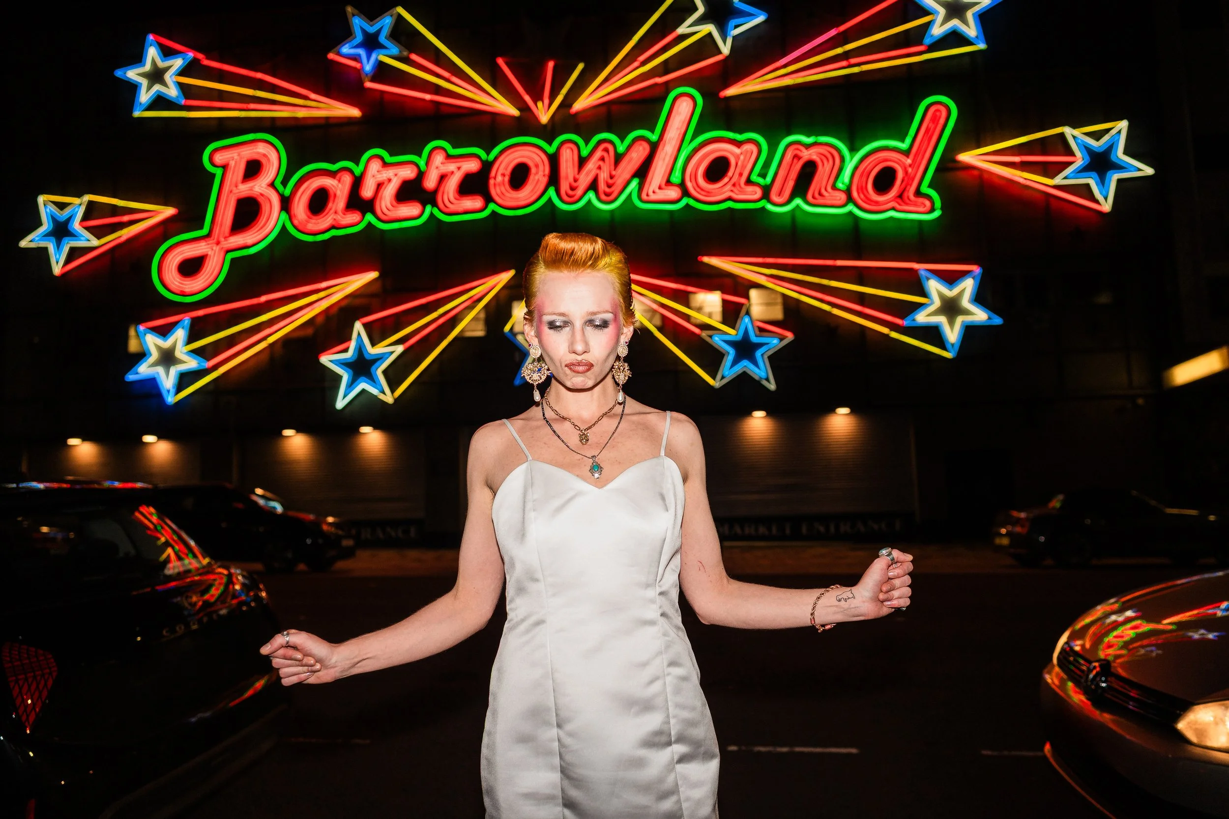 A woman in a white dress standing in front of a neon sign that says "Bazaland" with colorful star and streak decorations, at night with parked cars in the background.