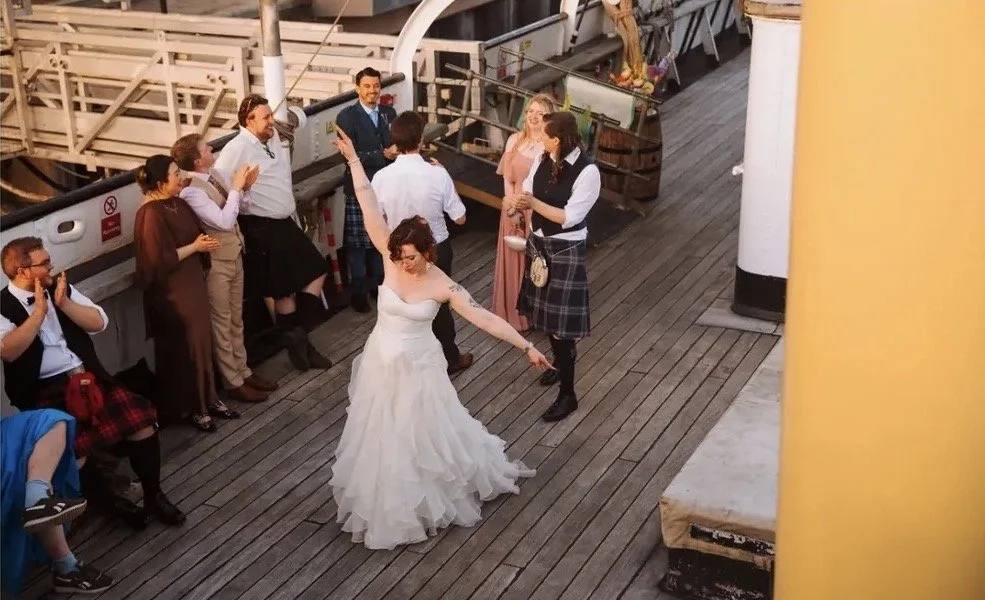 A bride dancing with her arms raised on the deck of a ship, surrounded by guests watching and clapping, dressed in formal attire.
