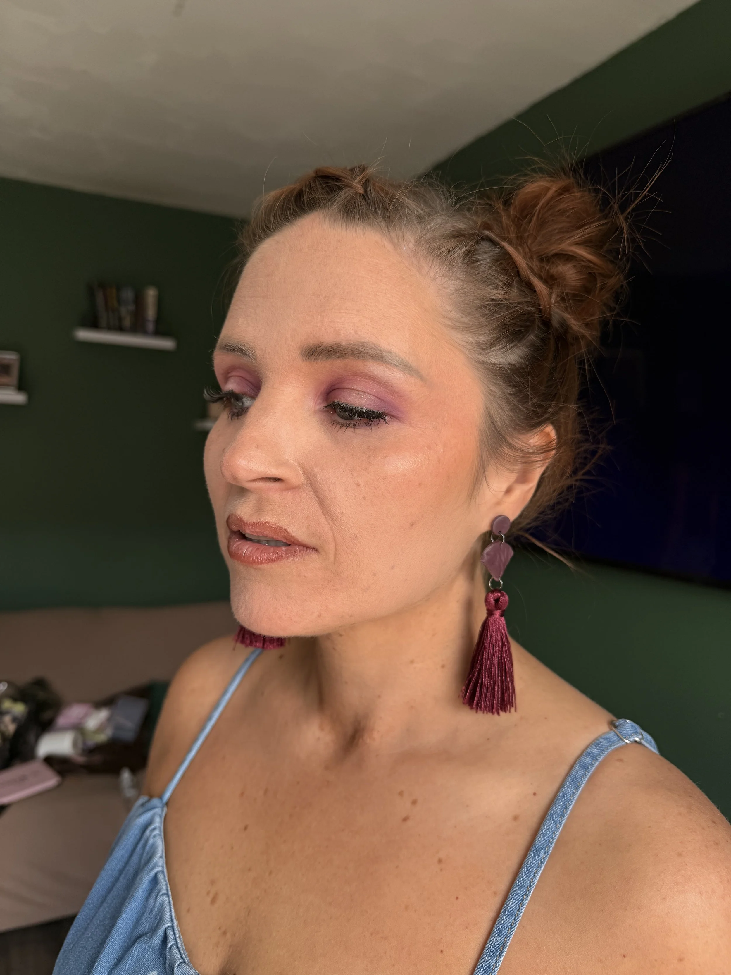 Close-up of a woman with reddish-brown hair styled in two space buns with loose strands, wearing purple and pink tassel earrings and a blue satin spaghetti strap top, with a green wall and shelves with books in the background.