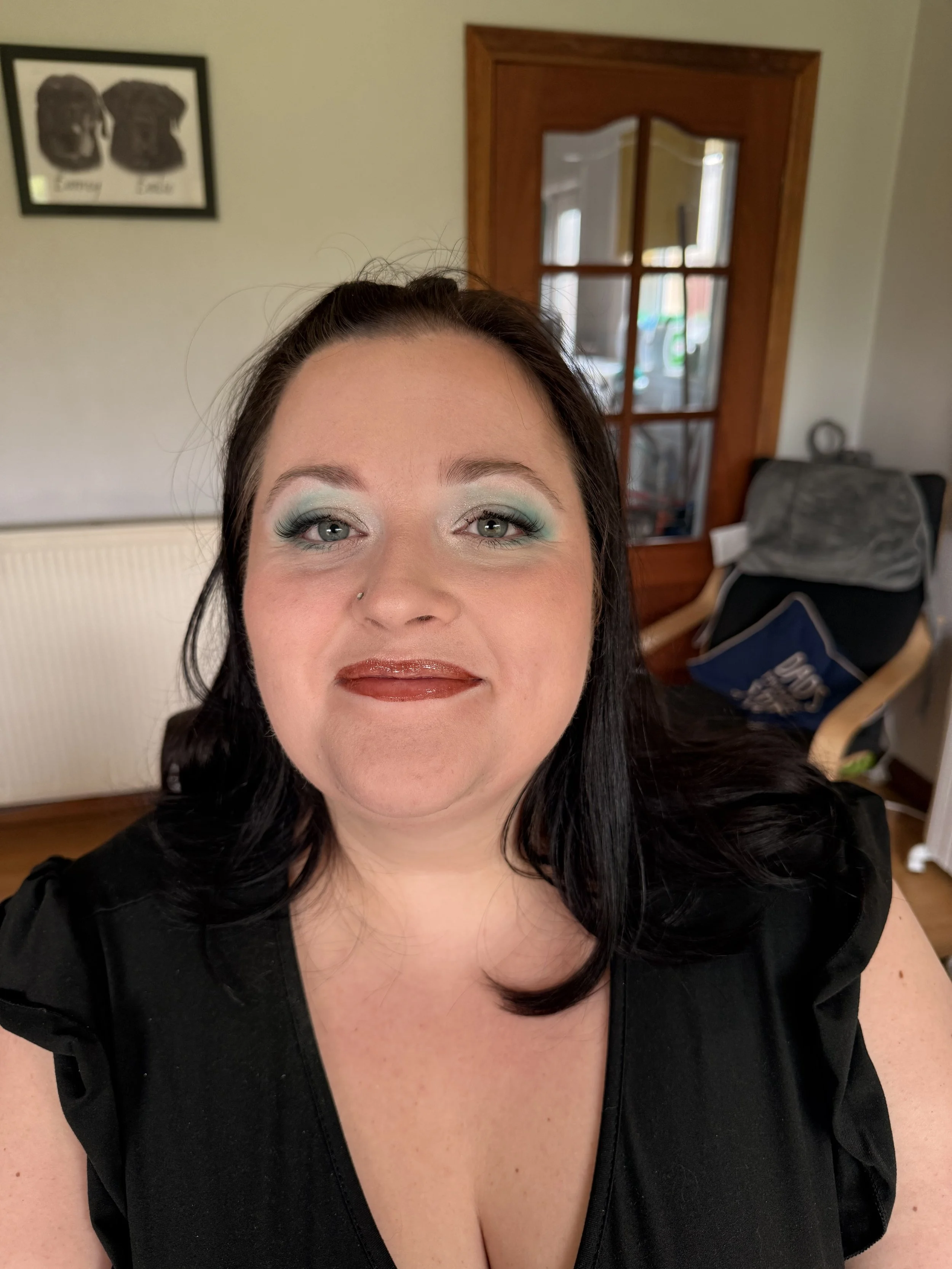 Close-up of a woman with dark hair, wearing makeup with turquoise eyeshadow and glossy lipstick, smiling indoors with a wooden door and framed pictures in the background.