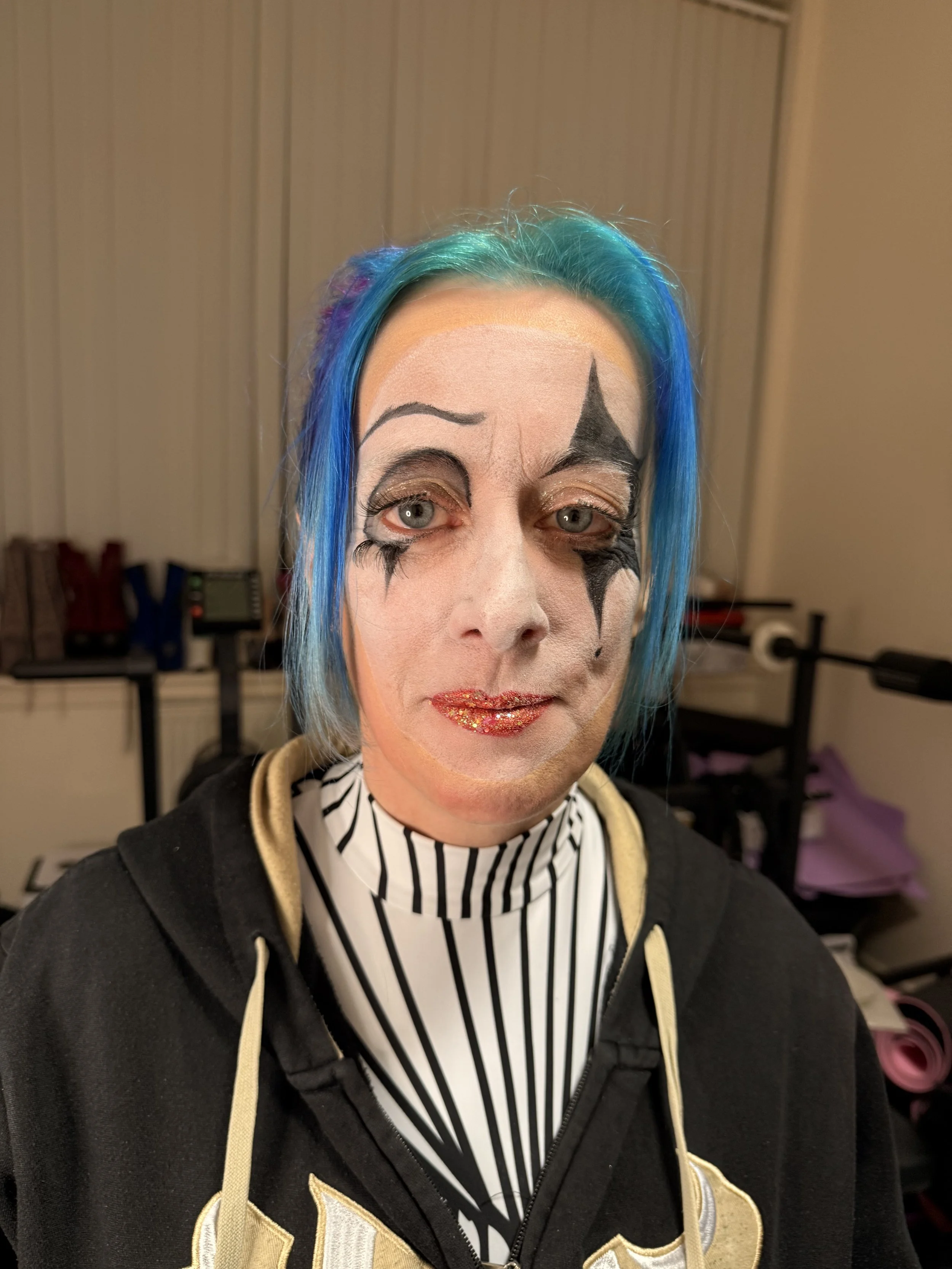 A person with blue hair and clown makeup, including black and white face paint, glittery lips, and artistic eye makeup, wearing a black hoodie with gold accents over a black and white striped shirt. The background shows a beige wall and some furniture.