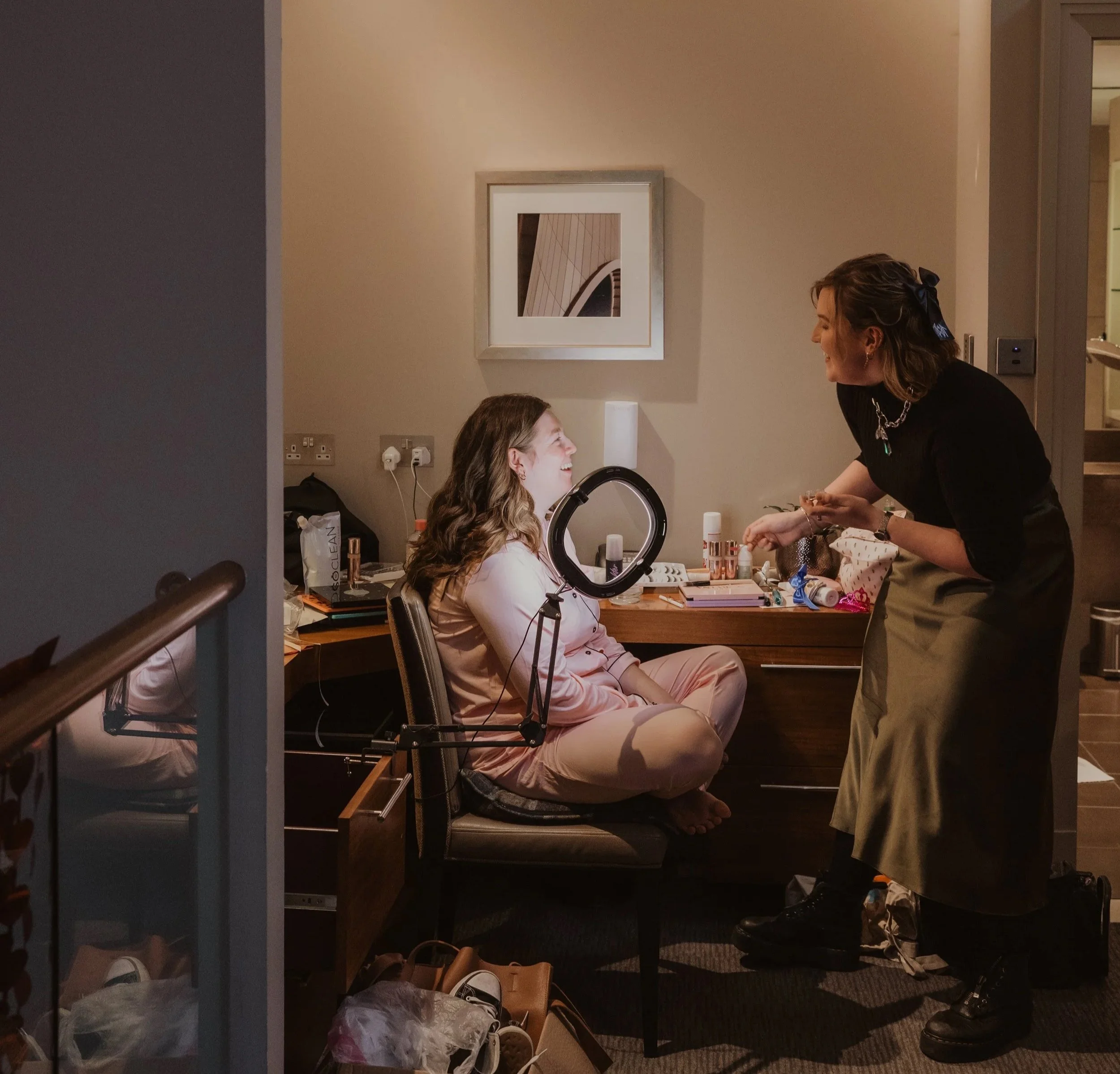 A woman with shoulder-length hair, wearing a dark top and a chain necklace, is smiling and leaning over a young girl with curly hair, who is sitting at a makeup table. The room has beige walls, a framed artwork, and a countertop with makeup and beauty products.