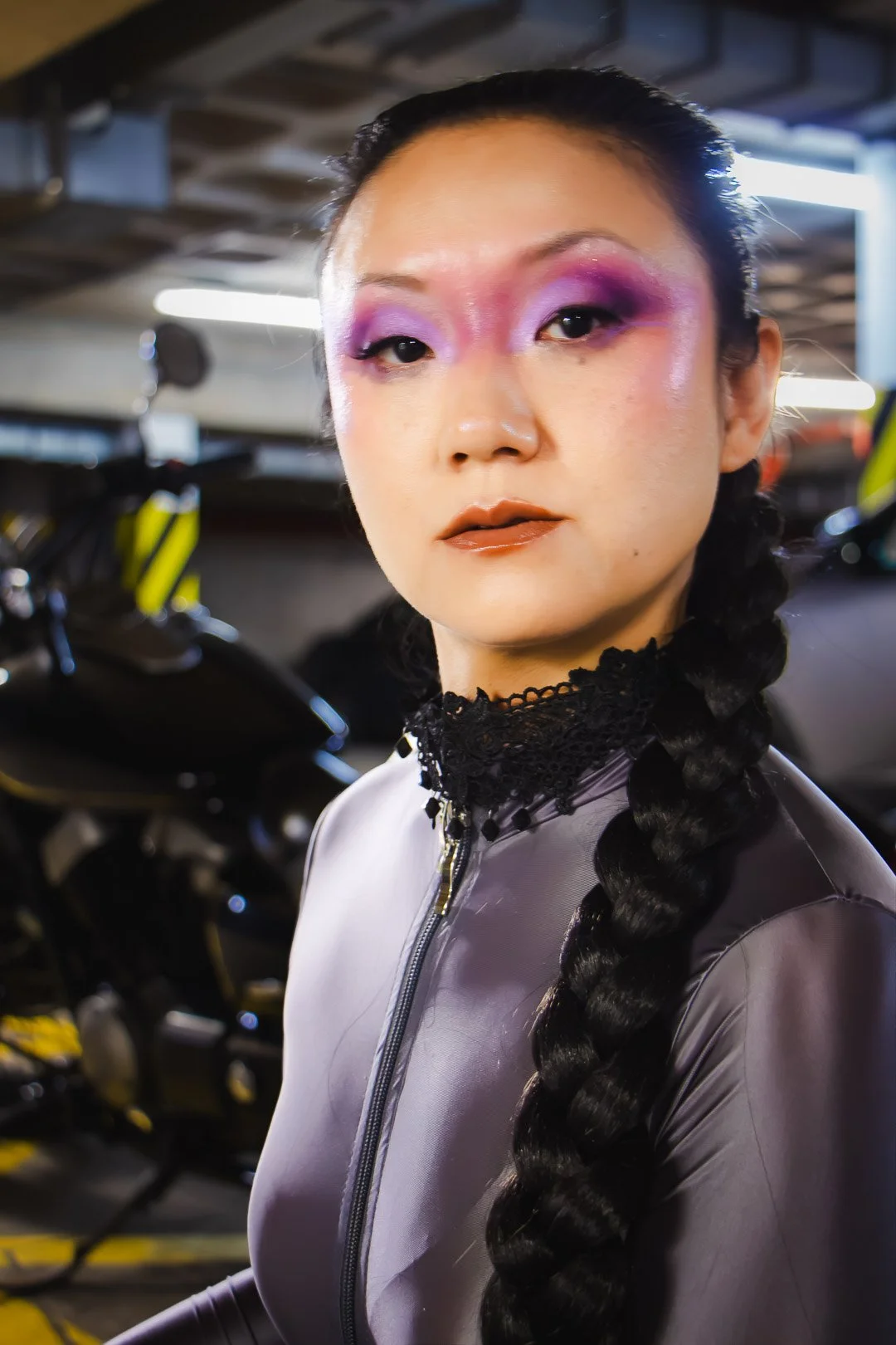 A woman with dramatic purple and pink eyeshadow, metallic lips, and a long braided hairstyle stands in a parking garage next to a black motorcycle.