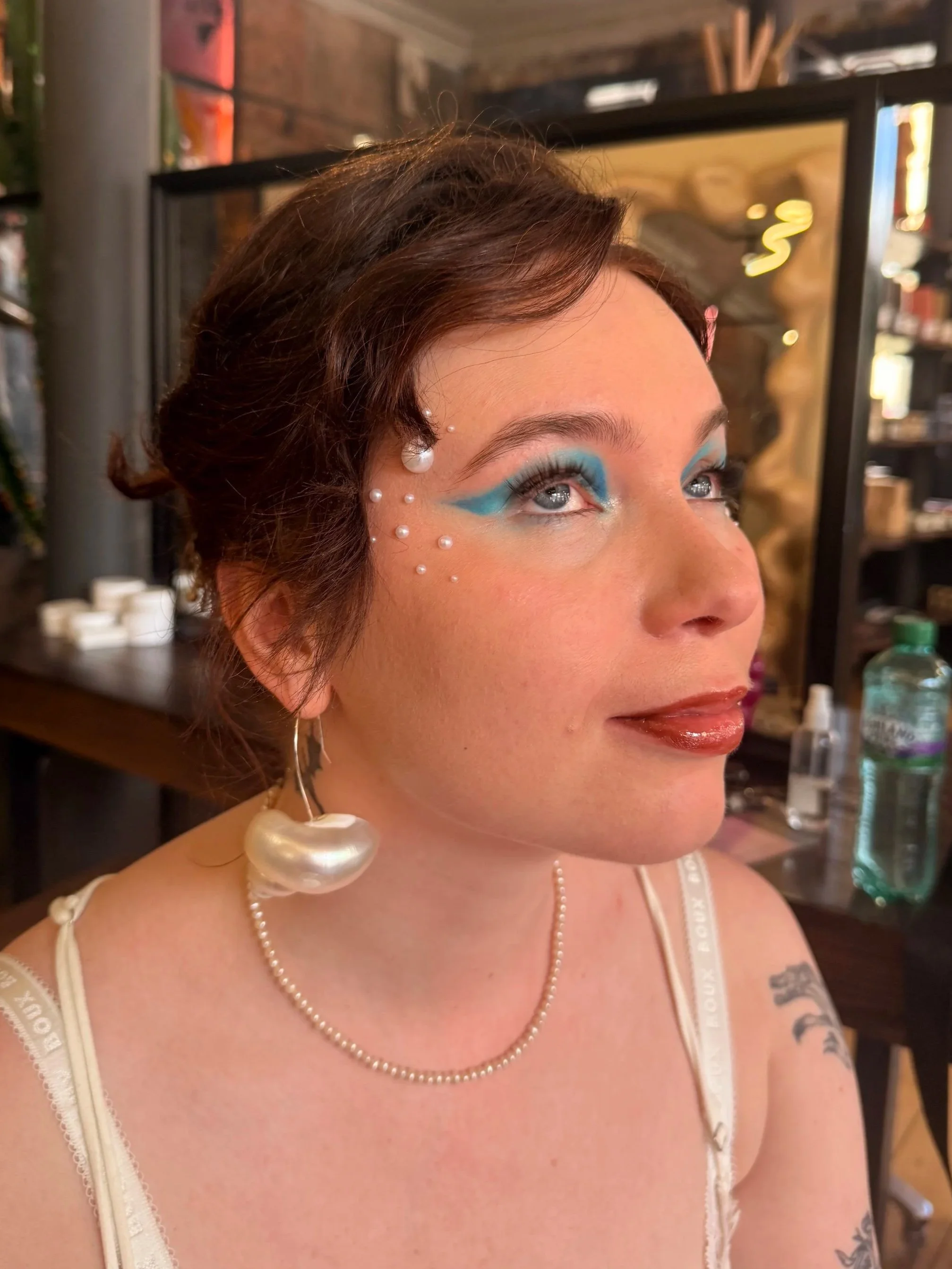 Close-up of a woman's face with blue eye makeup, pearl earrings, pearl necklace, and tattoo on her shoulder, indoors with shelves and bottles in the background.