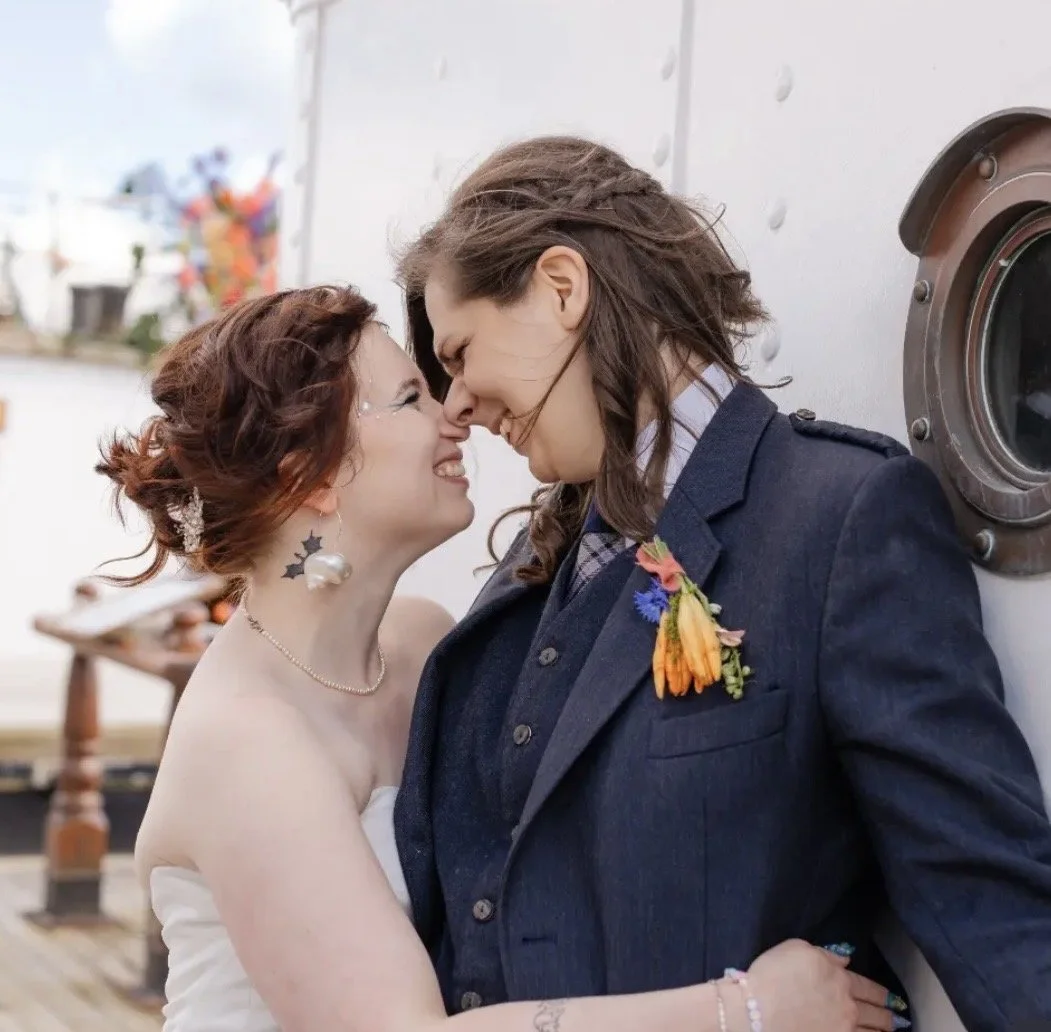 Two women with their foreheads touching and smiling, celebrating a wedding or special occasion, with festive decorations in the background.