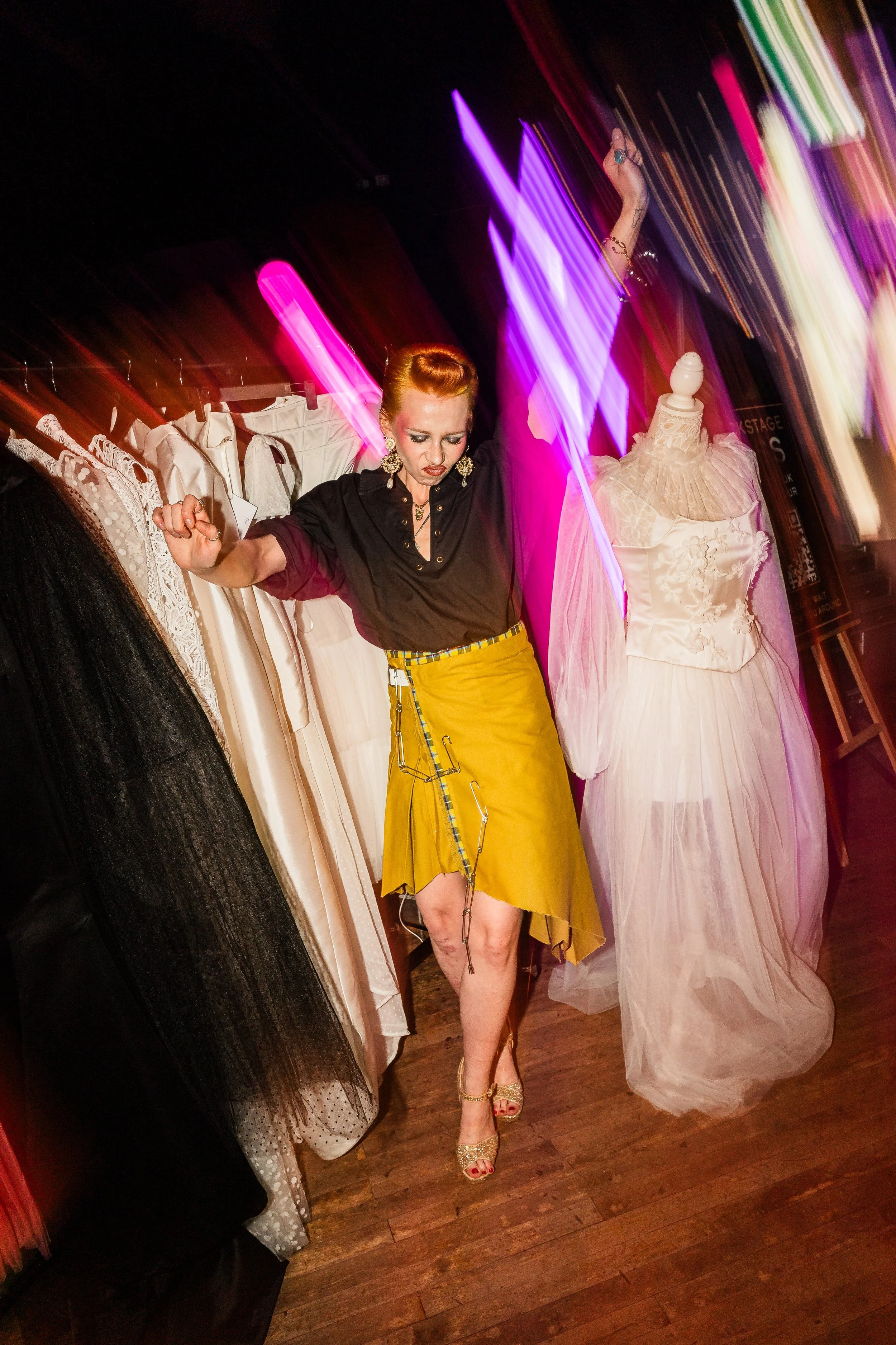A woman with red hair styled in an updo, wearing a black blouse and a yellow skirt with a chain detail, dancing in a room with dresses and mannequins; colorful lights create a blurred, dynamic background.