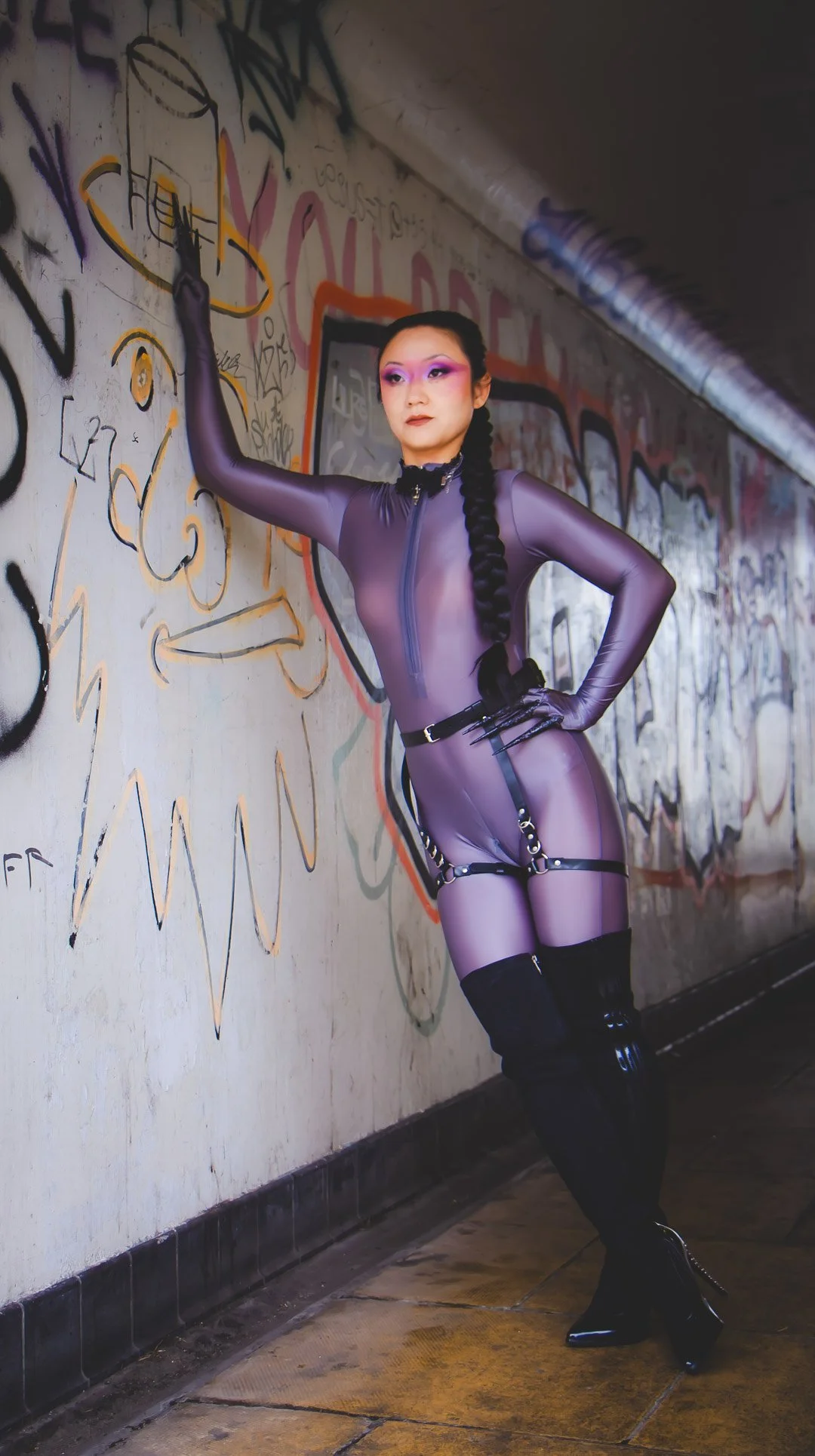 A person in a sheer purple bodysuit with black harness accessories, black thigh-high boots, and purple makeup, posing against a graffiti-covered wall in an urban setting.