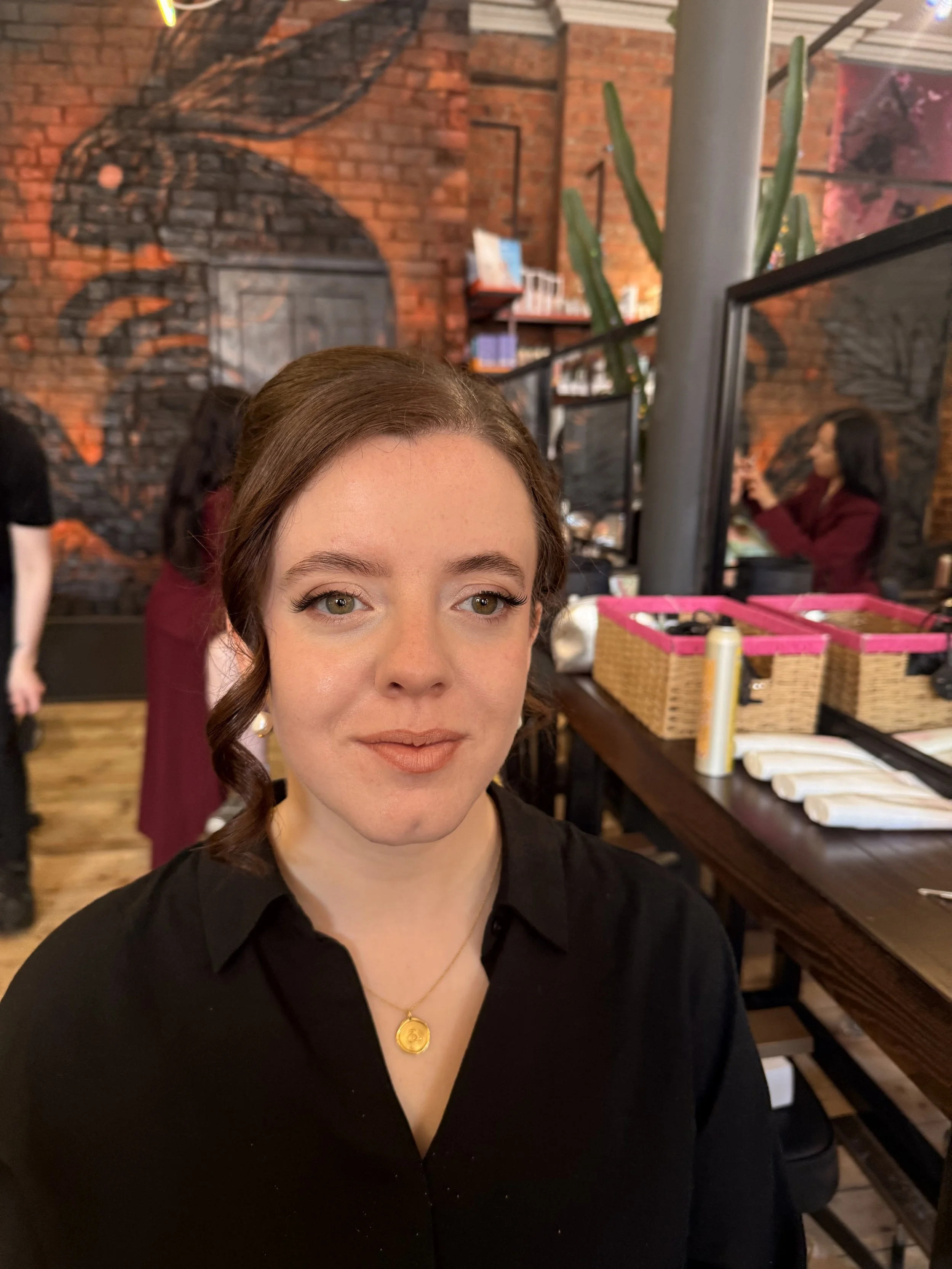 Close-up of a woman with brown hair styled in soft curls, wearing a black top, gold necklace, and earrings, in a cozy, stylish indoor setting with brick walls and artwork, with a woman in a maroon jacket sitting in the background and a wooden table w