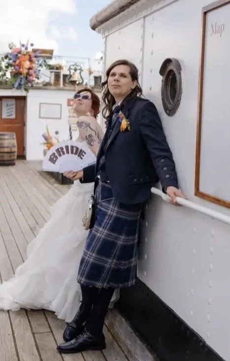 A woman in a plaid skirt and jacket leaning against a white wall on a boat deck, with a bride in a wedding dress and sunglasses holding a sign that says 'BRIDE' in the background.