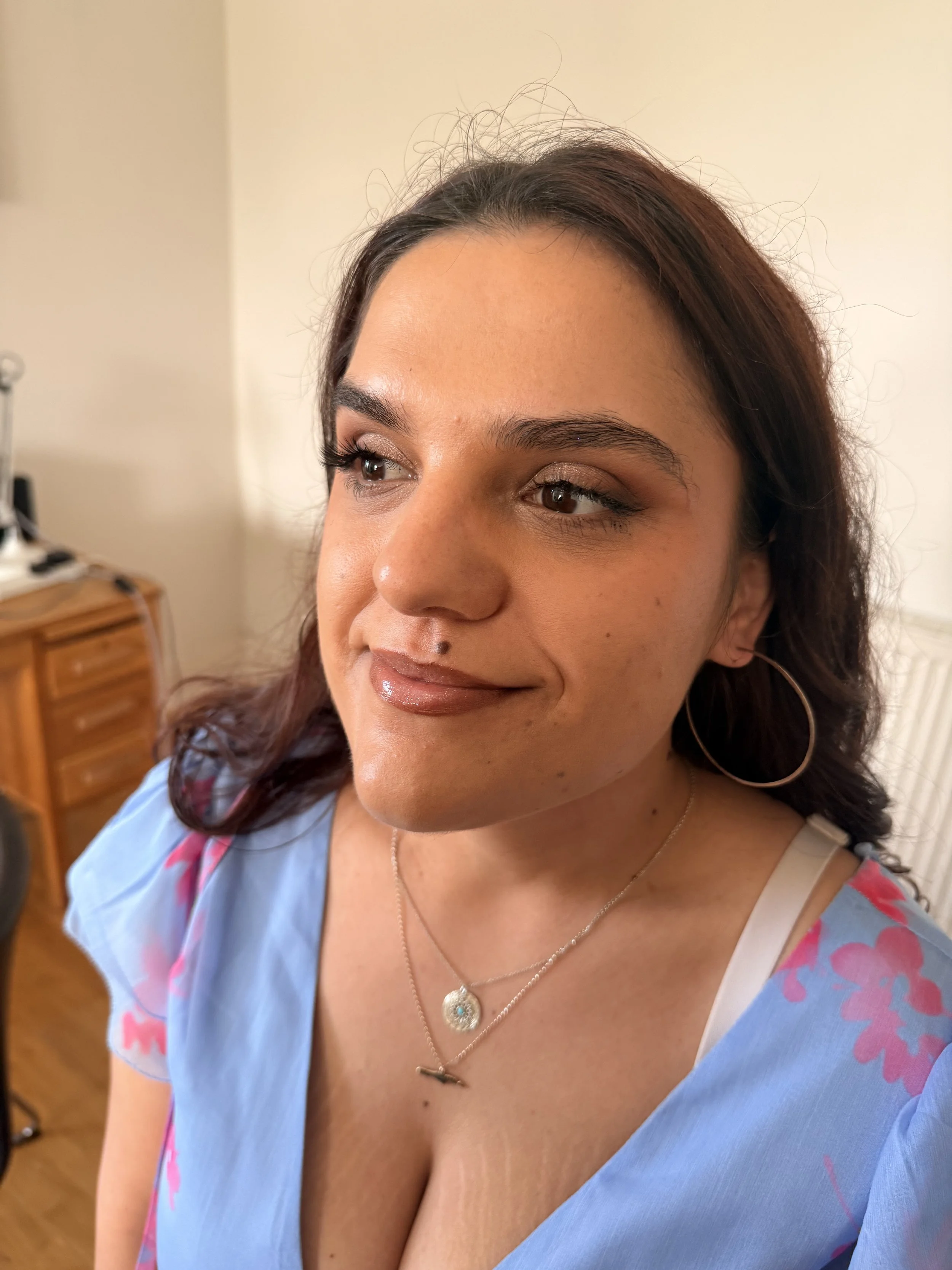 Close-up of a woman's face with makeup, wearing a blue top with pink floral pattern, gold hoop earrings, and layered necklaces, in a room with neutral-colored walls and a wooden dresser in the background.